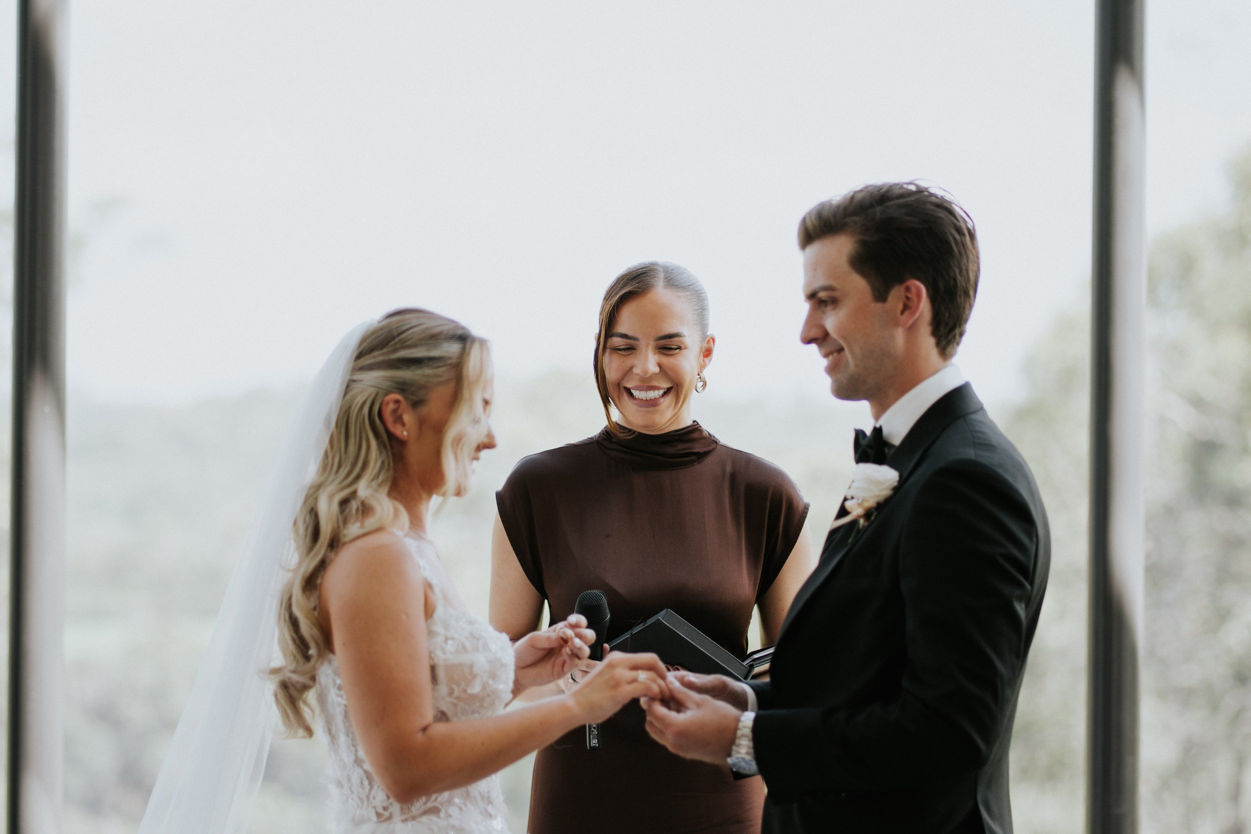 A wedding ceremony with a bride and groom exchanging vows, an officiant standing between them, captured in front of large windows with blurry outdoor scenery.