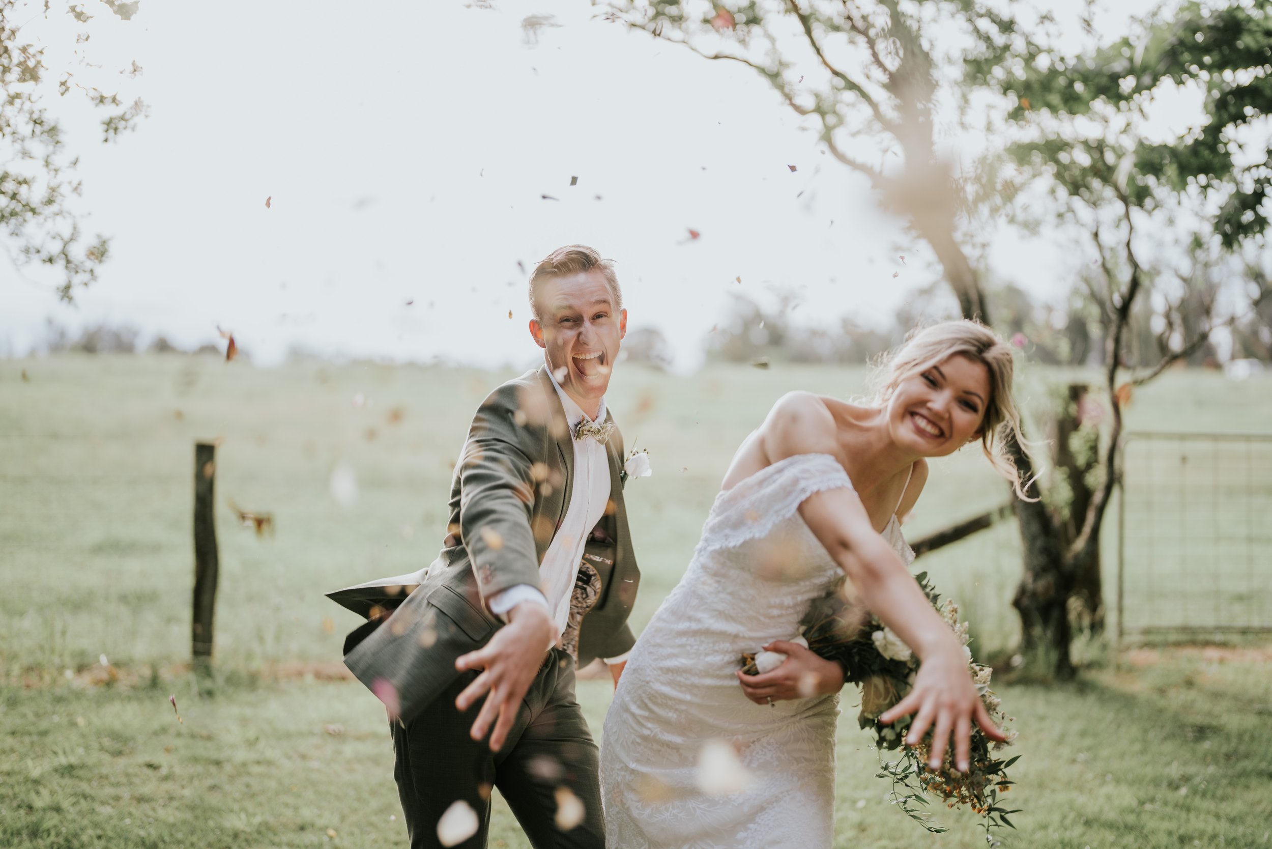 A newlywed couple in wedding attire celebrating outdoors, with the groom smiling and pointing at the camera and the bride smiling, holding a bouquet, as colorful confetti falls around them in a scenic, green park with trees and a fence.