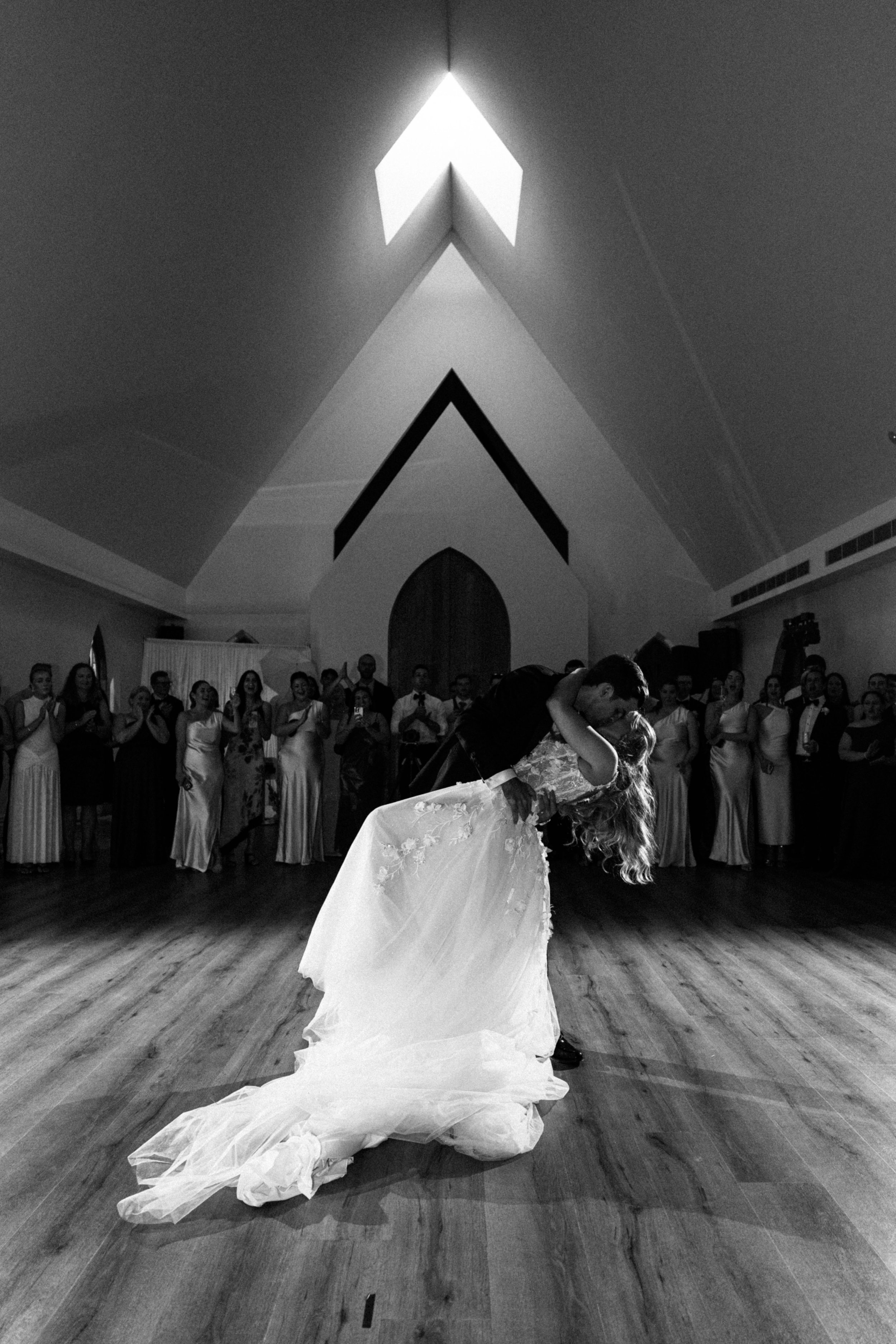 A bride and groom dance in front of a large audience in a wedding reception hall. The bride's long veil trails behind her as she is dipped by her groom during their dance. The ceiling has distinctive architectural features with a triangular light fix