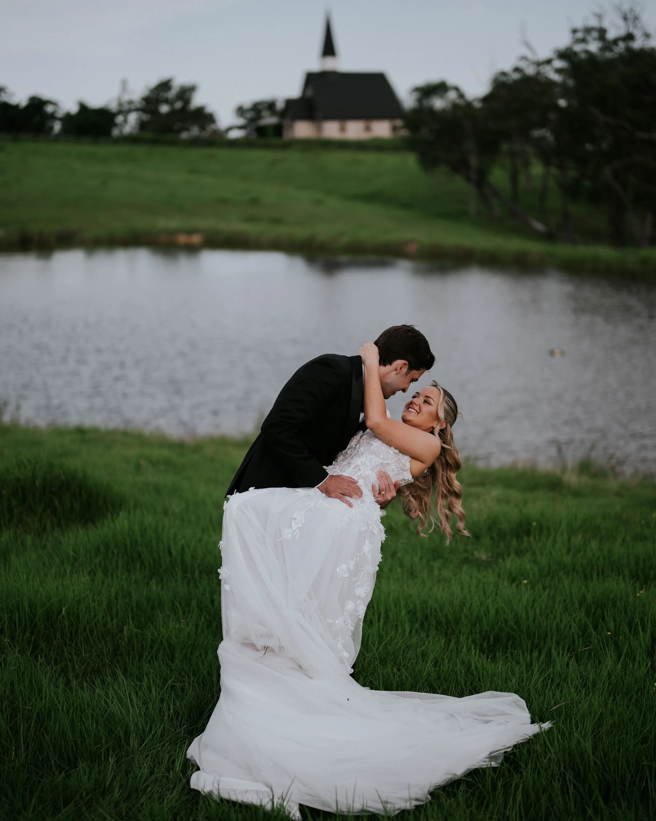 A newlywed couple embracing outdoors near a lake, with a church on a hill in the background, during early evening or dusk.