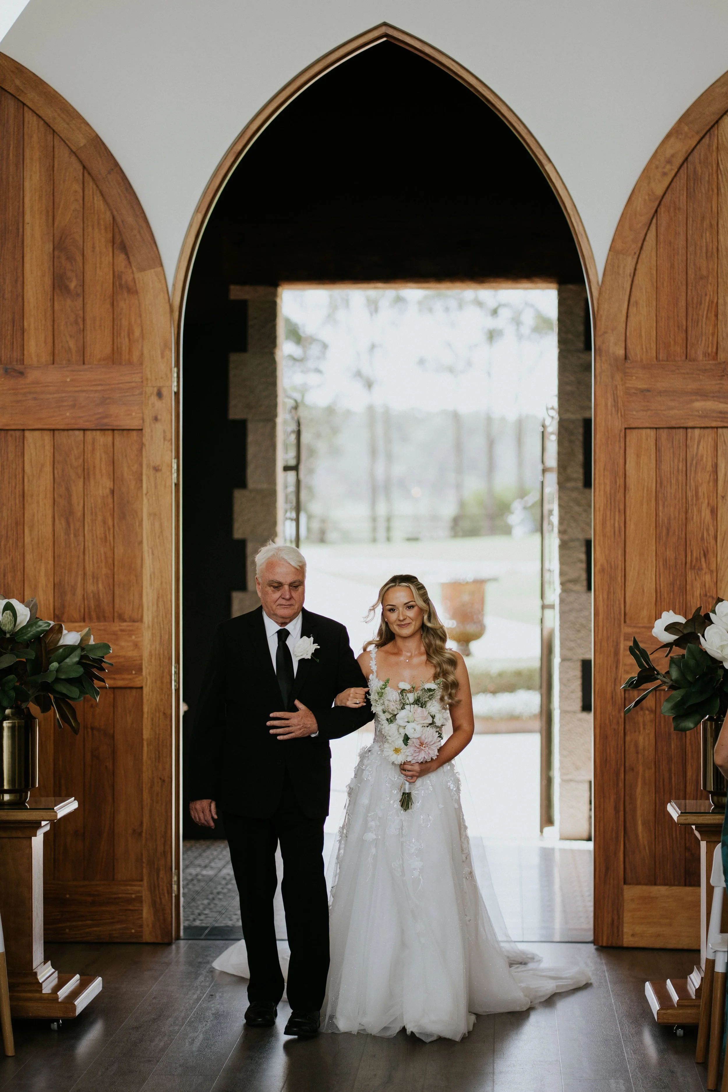 A bride in a white wedding gown holding a bouquet of white and pink flowers walks down the aisle with an older man, possibly her father, inside a wooden church with large open doors and natural light coming from outside.