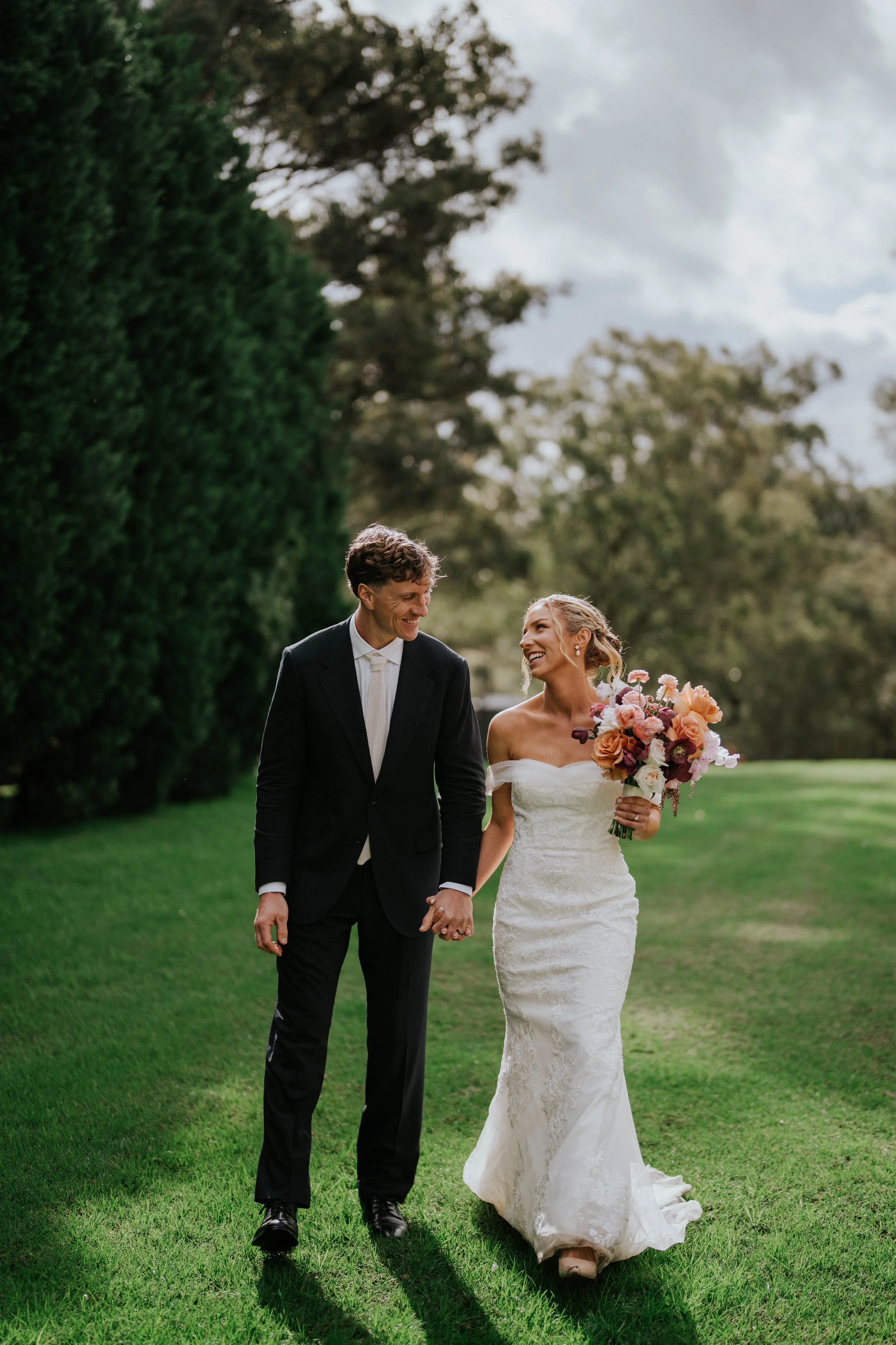 A bride and groom walking hand in hand on a lush green lawn, smiling at each other during their wedding celebration, with trees and a cloudy sky in the background.