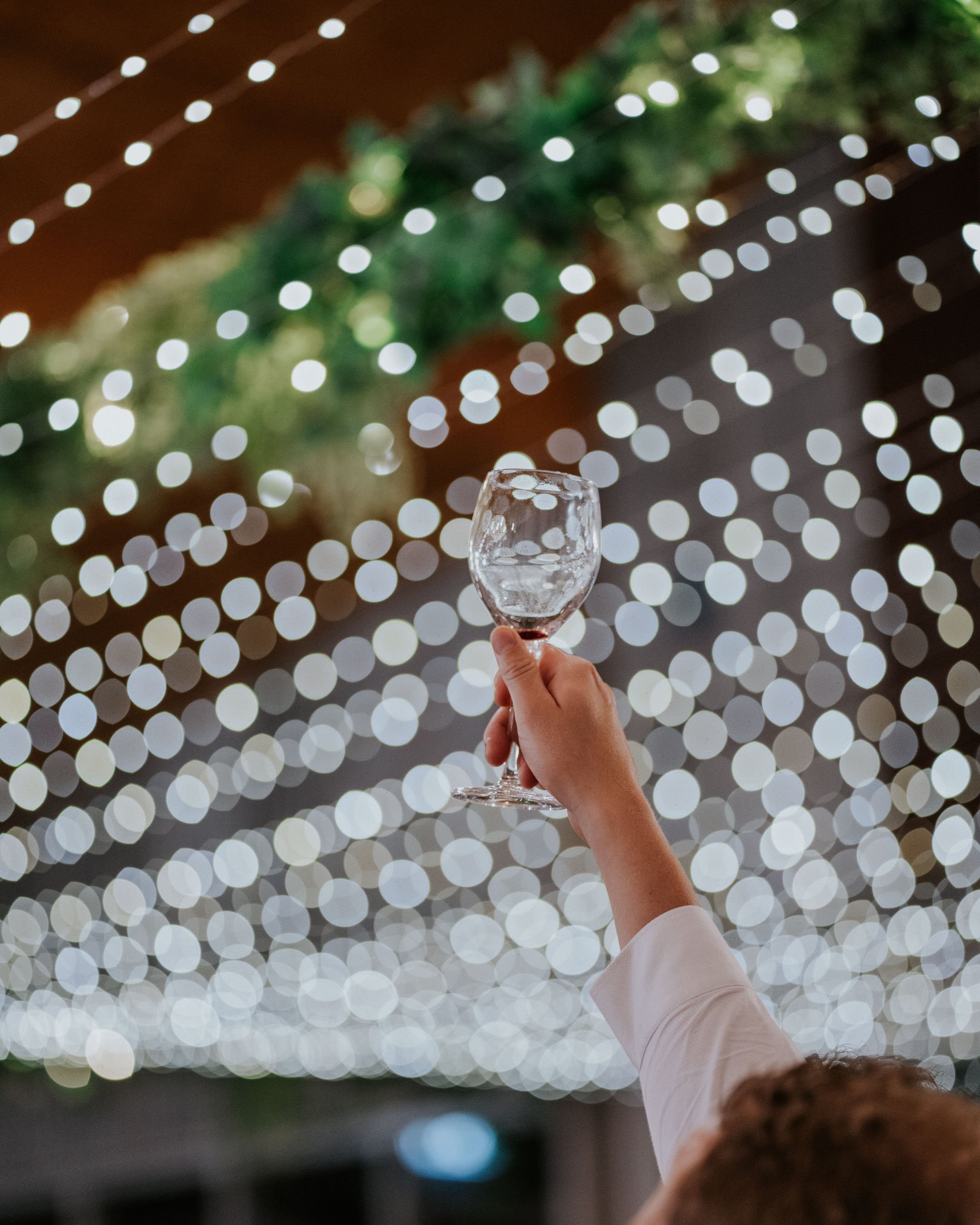 A hand holding an empty wine glass raised in a toast, against a background of blurred string lights and greenery, likely at a festive outdoor event.