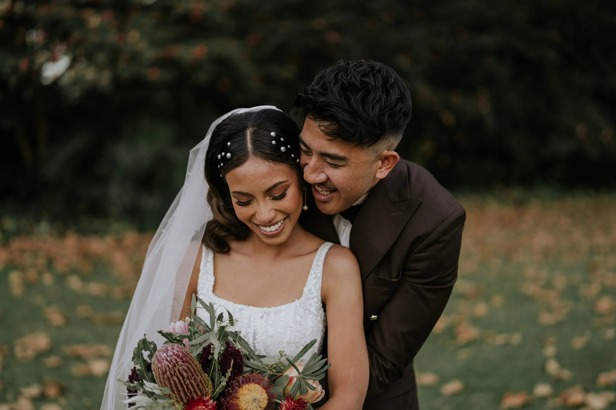 A bride and groom smiling, embracing outdoors during their wedding. The bride is holding a bouquet of flowers with pink, red, and yellow blooms, and the groom is wearing a dark suit. The background is blurred greenery.