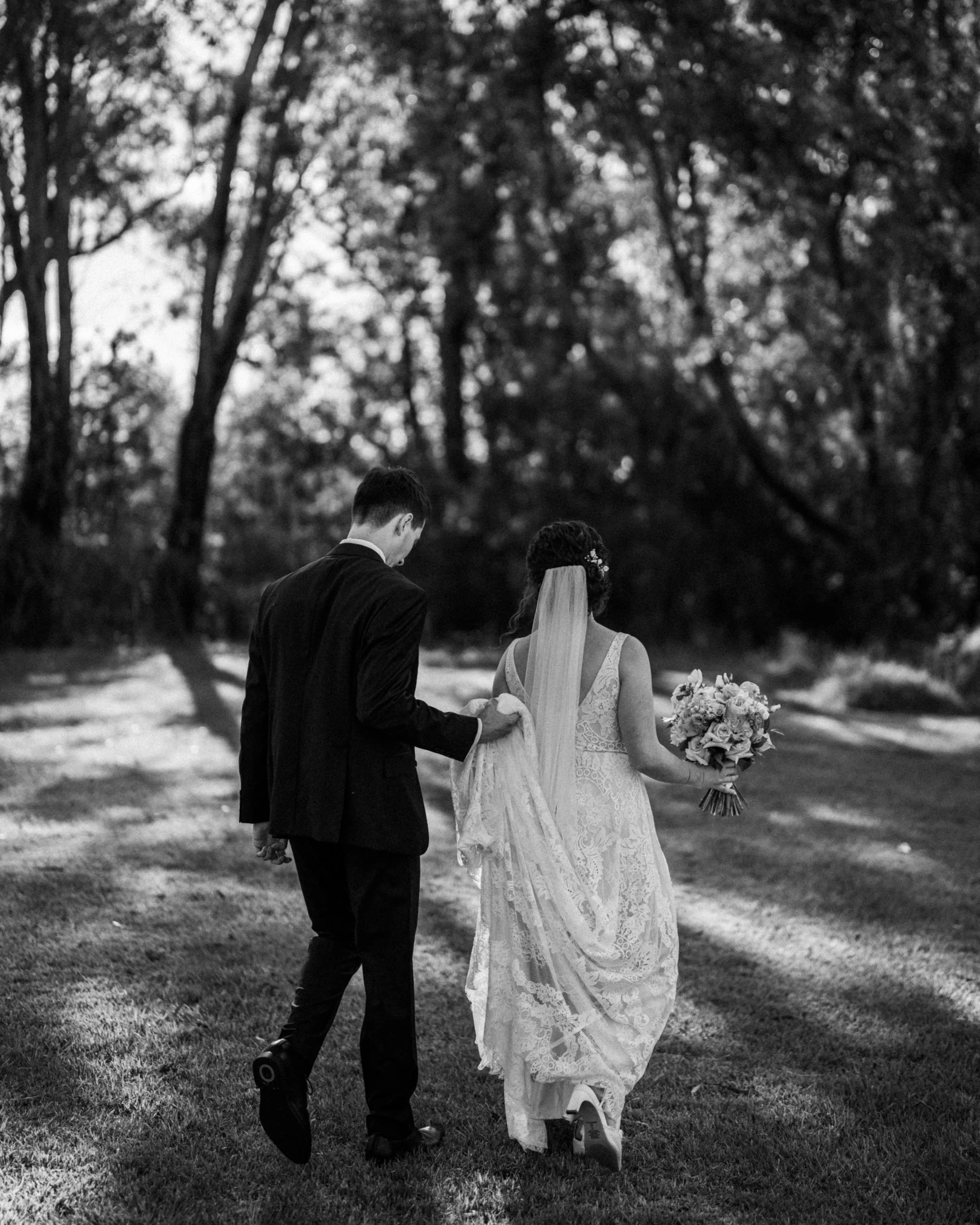 This black and white photo depicts a bride and groom walking together outdoors on grass. The groom is dressed in a dark suit, and the bride wears a white lace wedding dress and holds a bouquet of flowers. They are walking away from the camera with their heads close together, surrounded by trees.