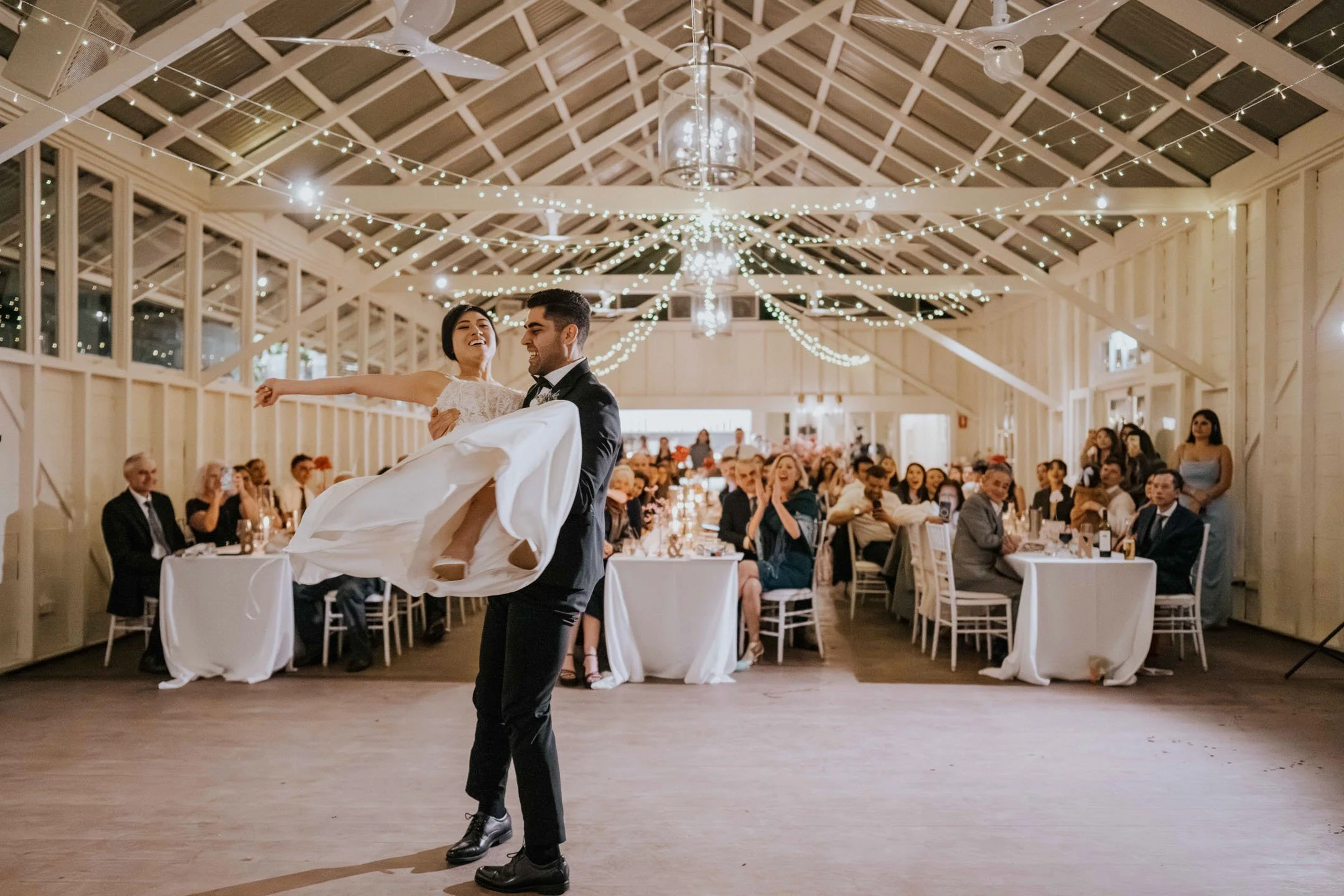 A groom in a black tuxedo holding a bride in a white wedding dress during their first dance at a wedding reception, with guests seated at tables watching in a decorated hall with string lights on the ceiling.