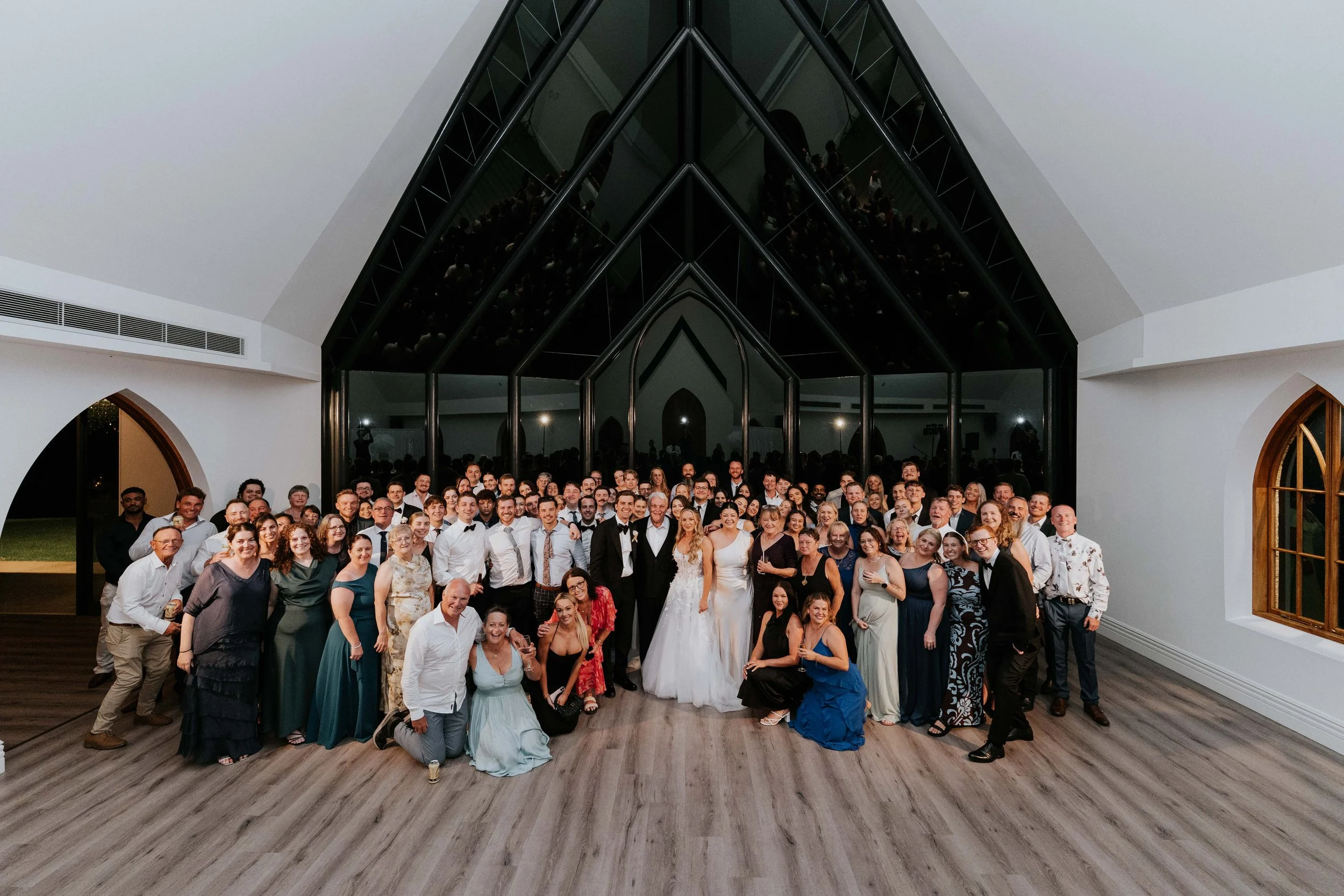 Large group of people at wedding reception, with a bride and groom in wedding attire at the center, inside a modern venue with glass walls.