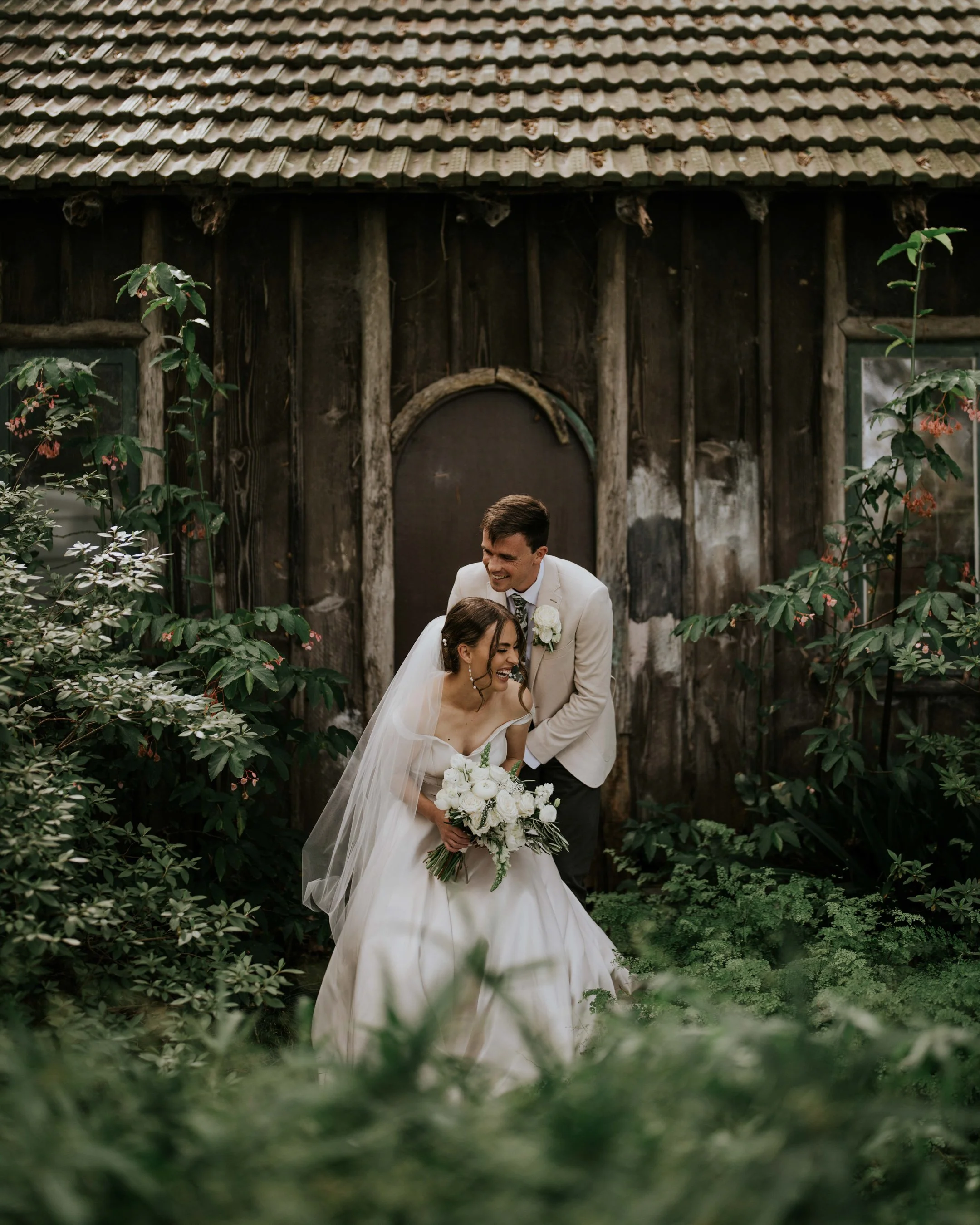 A bride and groom smiling and laughing during their wedding ceremony or photo shoot outdoors, surrounded by green foliage, in front of a rustic wooden building with a tiled roof.