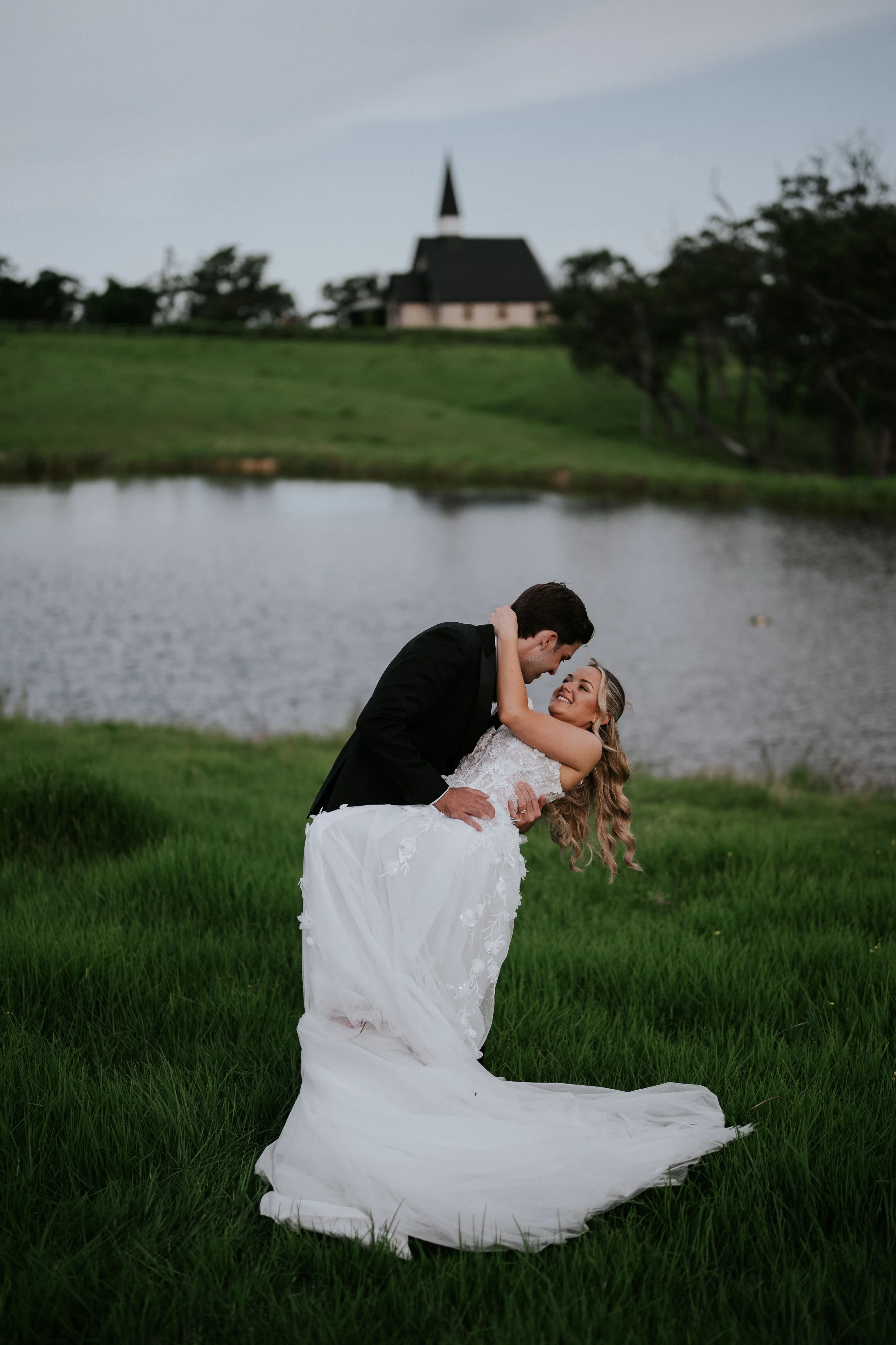 A bride and groom are dancing outdoors near a body of water with a church on a hill in the background on a grassy area.