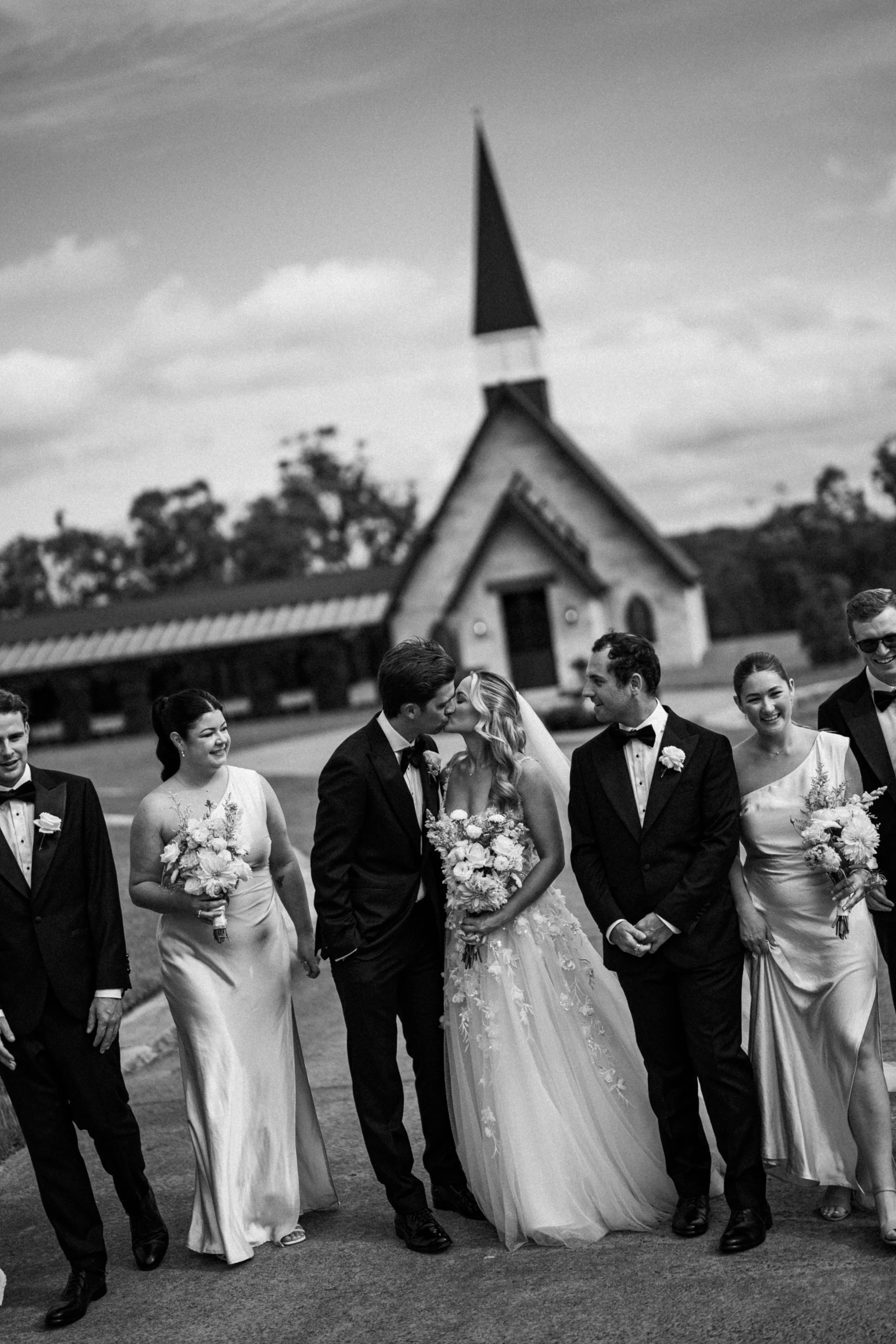 Black and white photo of a wedding party outdoors, with a church in the background. The bride and groom are kissing, surrounded by bridesmaids and groomsmen holding bouquets and wearing formal attire.