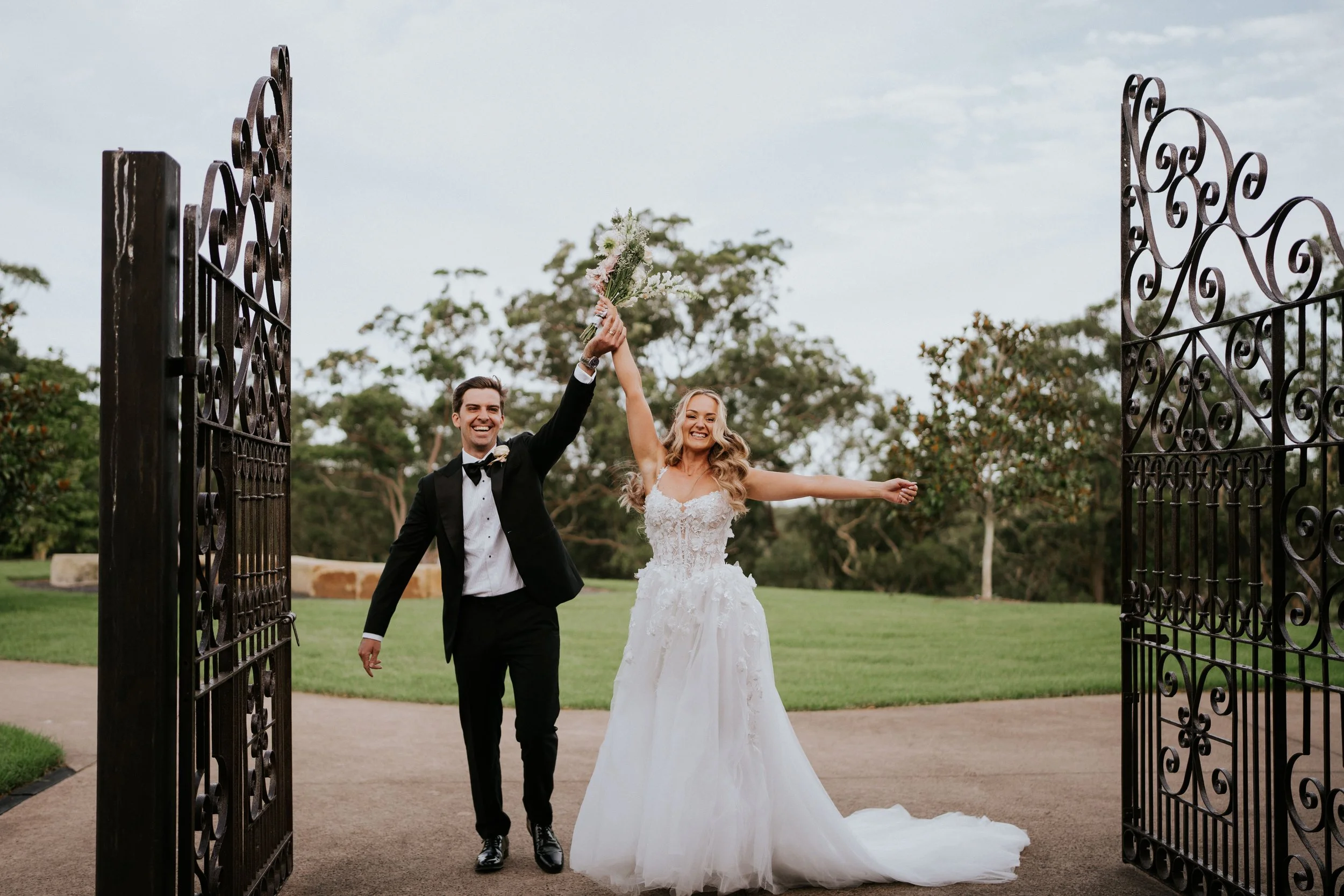 A newlywed couple at their wedding, the groom in a black tuxedo and the bride in a white wedding dress, holding hands through an open wrought iron gate, smiling and celebrating outdoors.