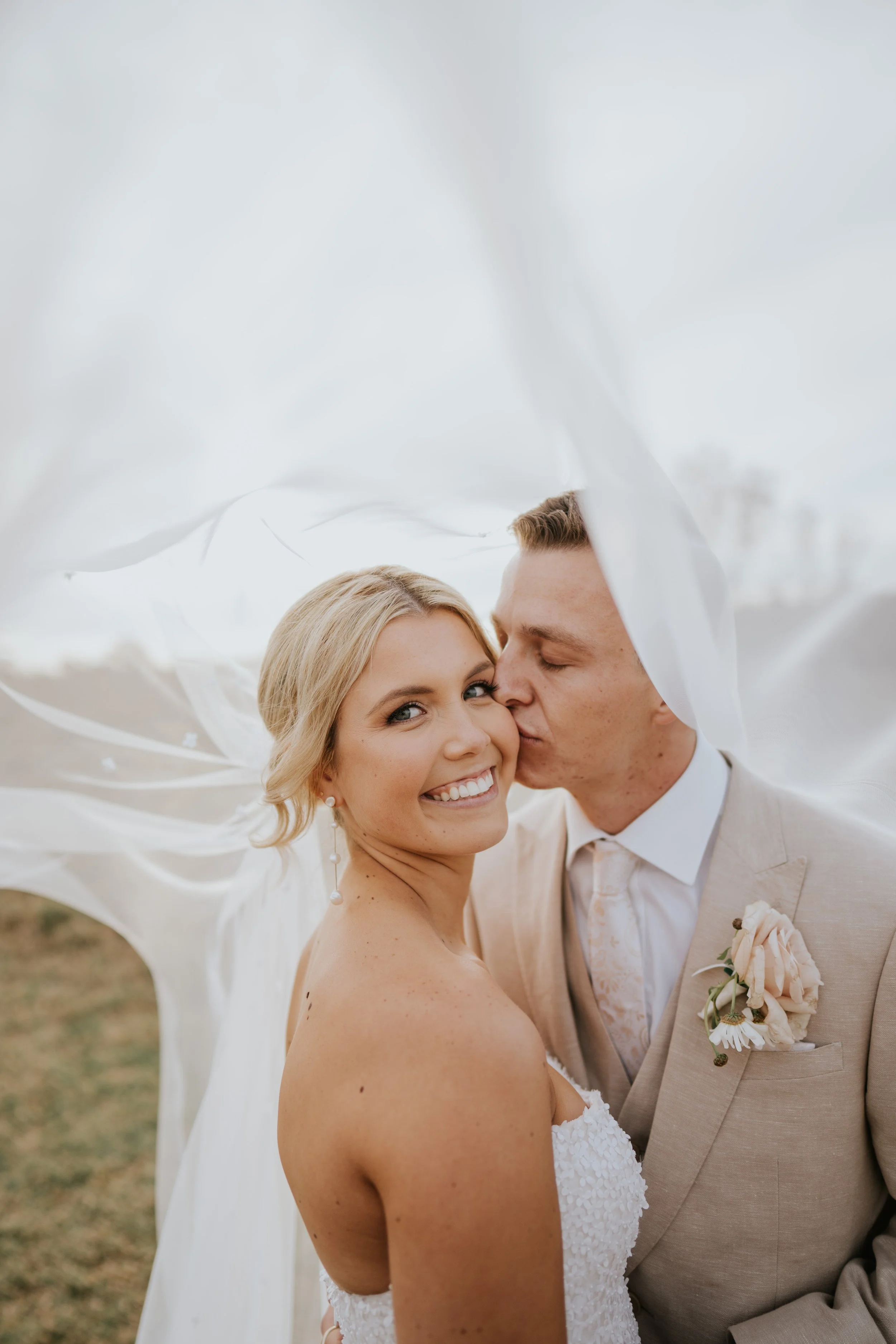 A bride and groom sharing an intimate moment under a flowing white fabric, outdoors, during their wedding, with the bride smiling and looking at the camera, and the groom kissing her on the cheek.