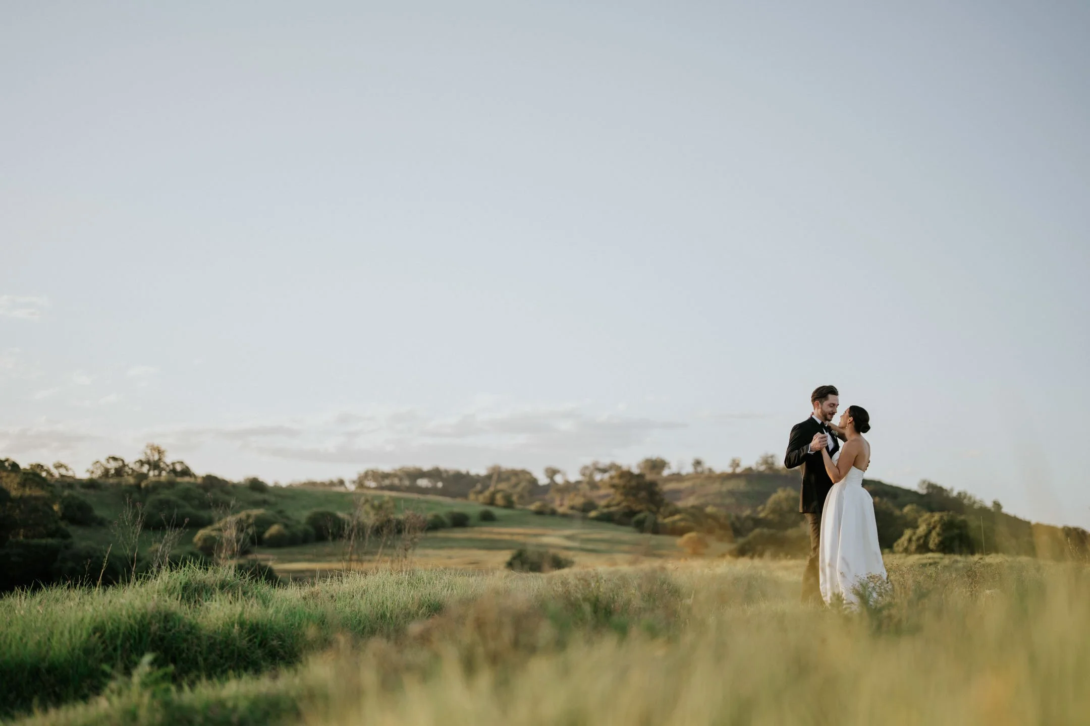 A newlywed couple dancing outdoors in a grassy field with hills and trees in the background during sunset or late afternoon.