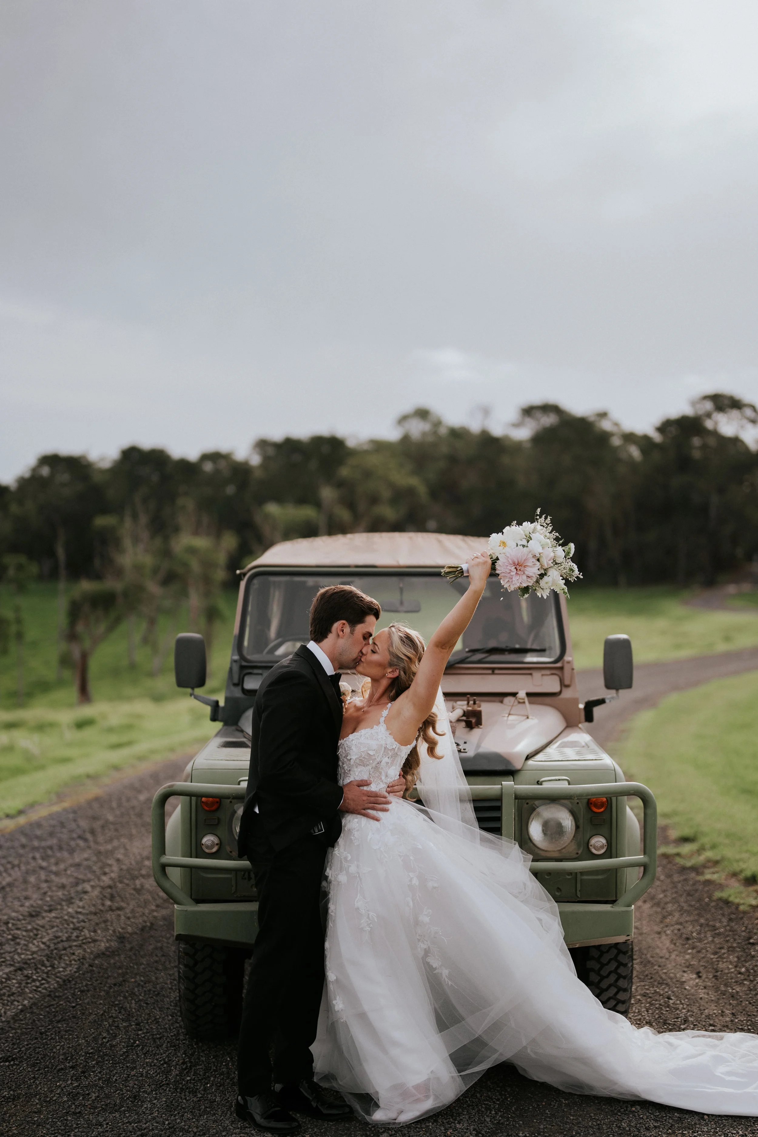 A bride and groom share a kiss in front of a vintage off-road vehicle during a outdoor wedding photoshoot. The bride is holding a bouquet and wearing a white wedding dress, the groom is in a black tuxedo. The background features a rural landscape wit