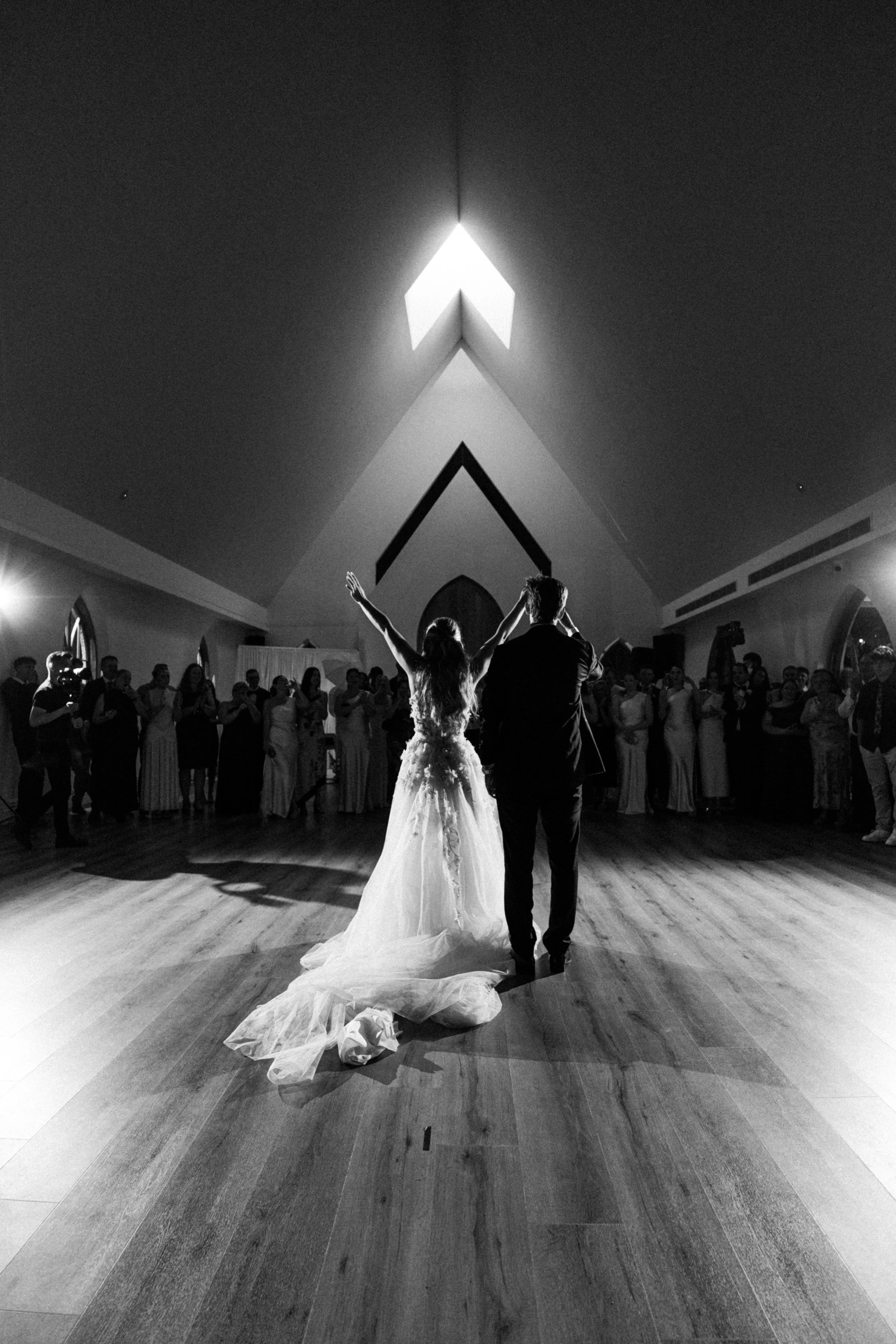 Black and white photo of a bride and groom dancing in a church or reception hall, with guests watching in the background. The bride wears a long gown with a train, and the groom is dressed in a suit. The ceiling has a triangular window letting light 