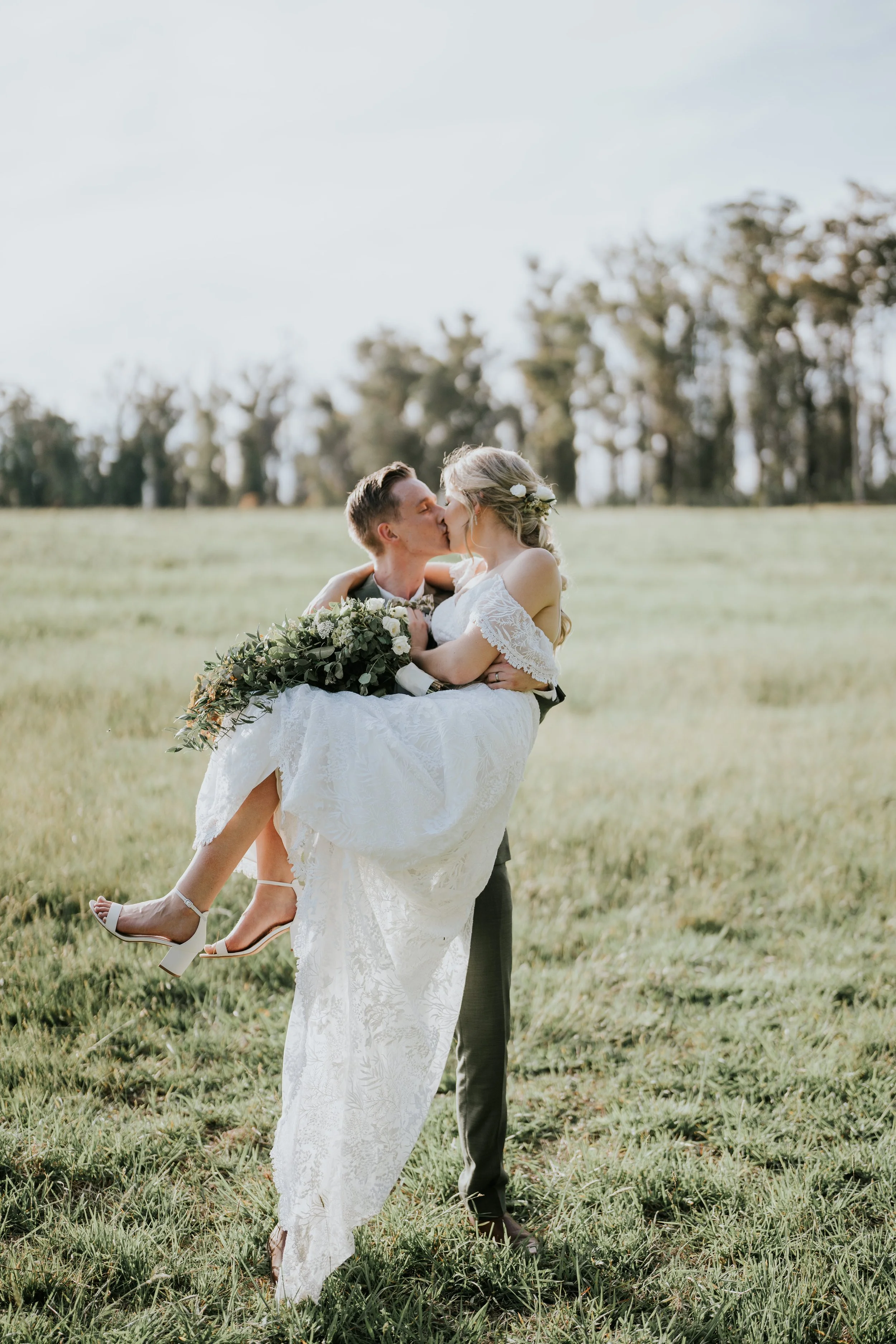 A groom lifts a bride in a white lace wedding dress in a grassy field during daytime, kissing and holding her.