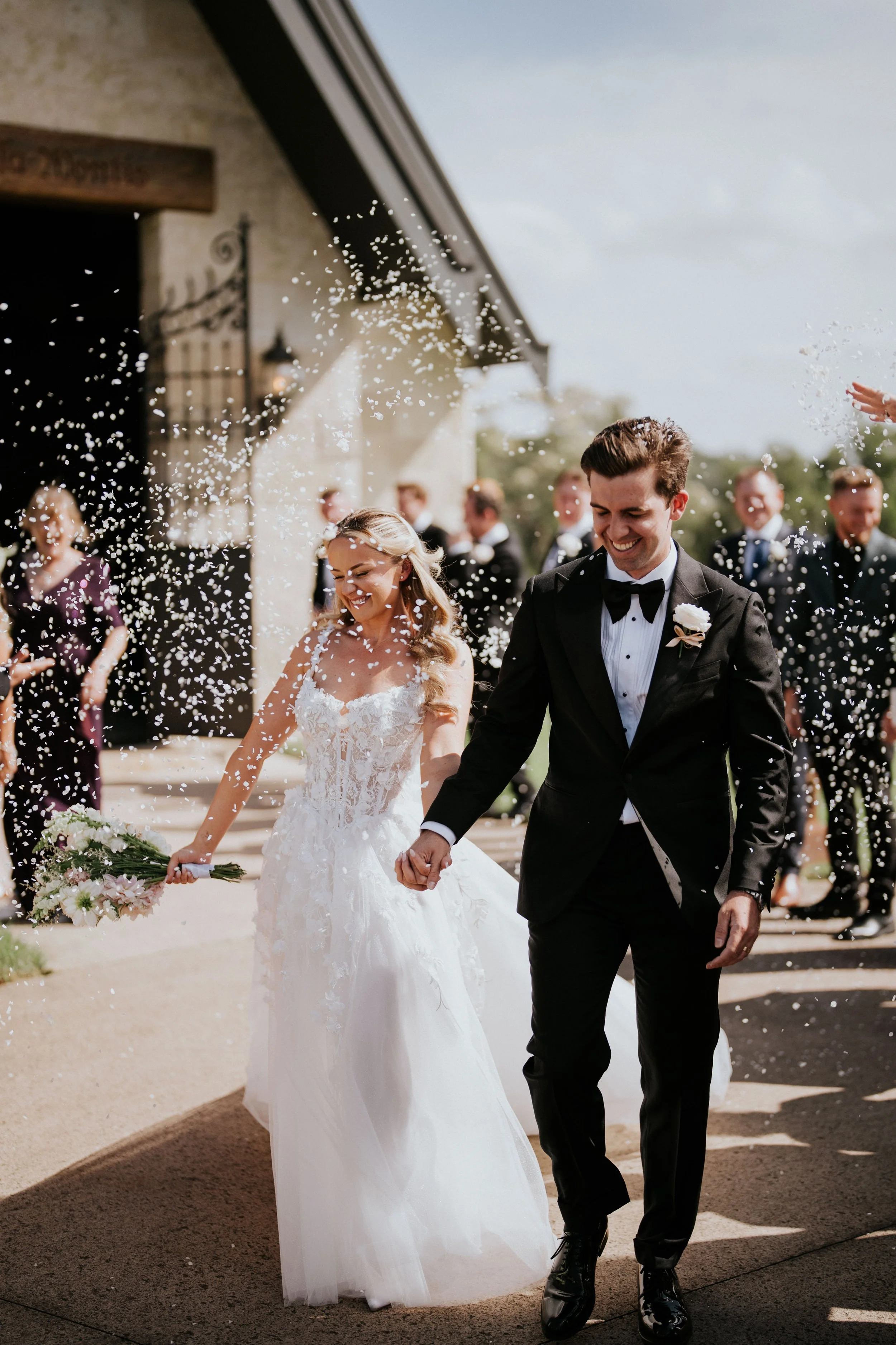 A newlywed couple walking hand in hand, smiling and surrounded by people celebrating outside during daytime. The bride wears a white wedding gown and holds a bouquet, and the groom is in a black tuxedo with a bow tie. Confetti is falling around them.