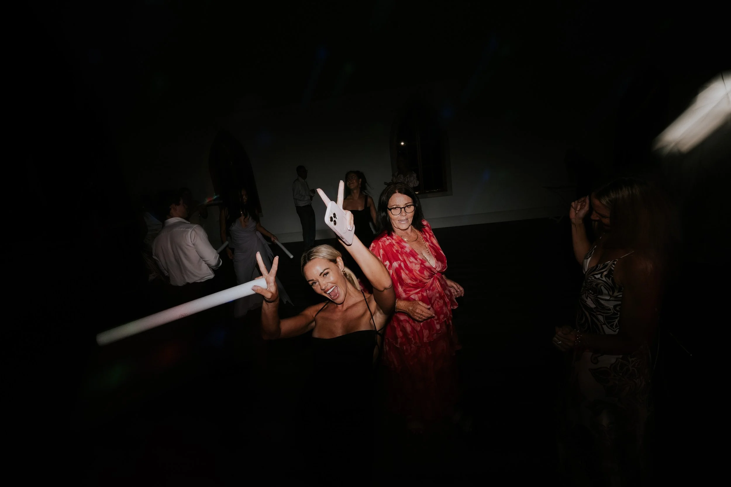 People at a party, some smiling and taking selfies, in a dimly lit room.