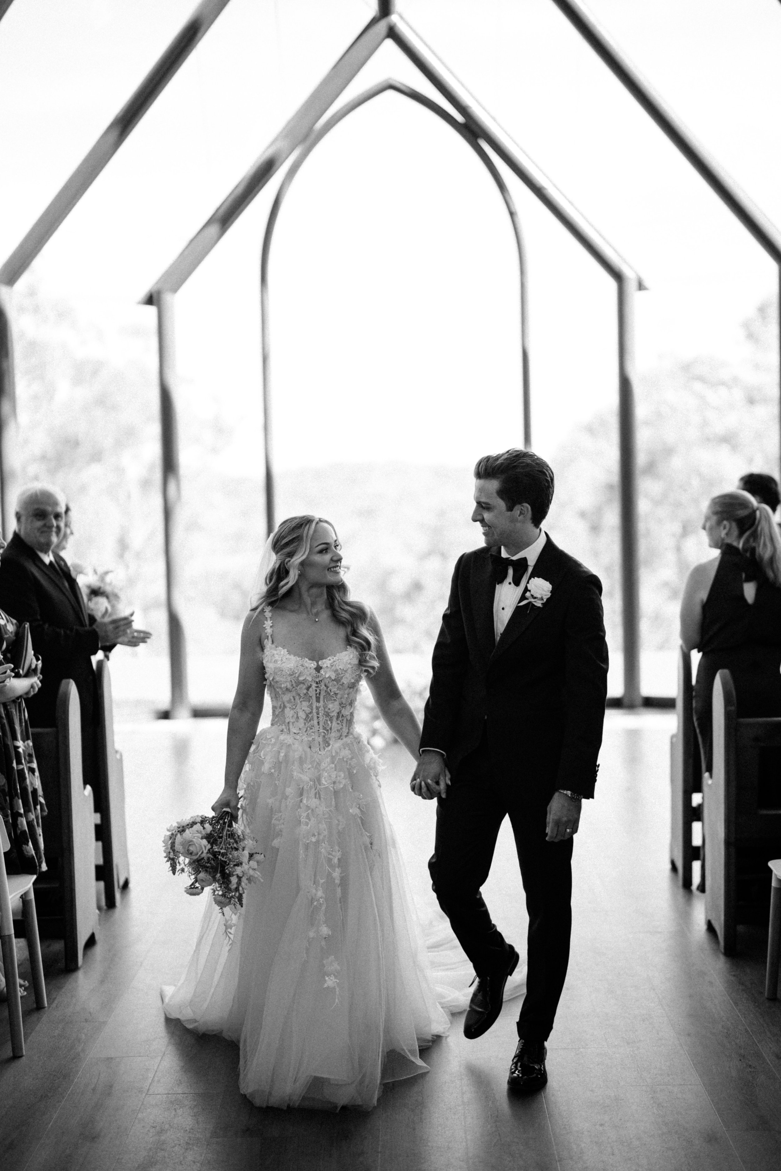A black and white photo of a bride and groom walking hand in hand down the aisle after their wedding ceremony. The bride is wearing a lace wedding dress and holding a bouquet, while the groom is dressed in a tuxedo. Guests are standing on either side
