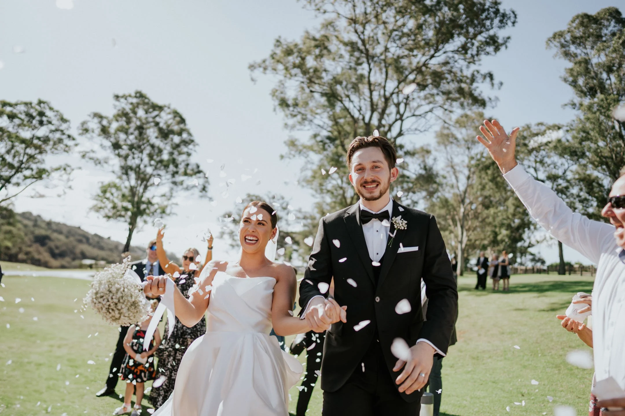 A joyful bride and groom walk hand in hand through a confetti shower outdoors, surrounded by friends and family celebrating their wedding.