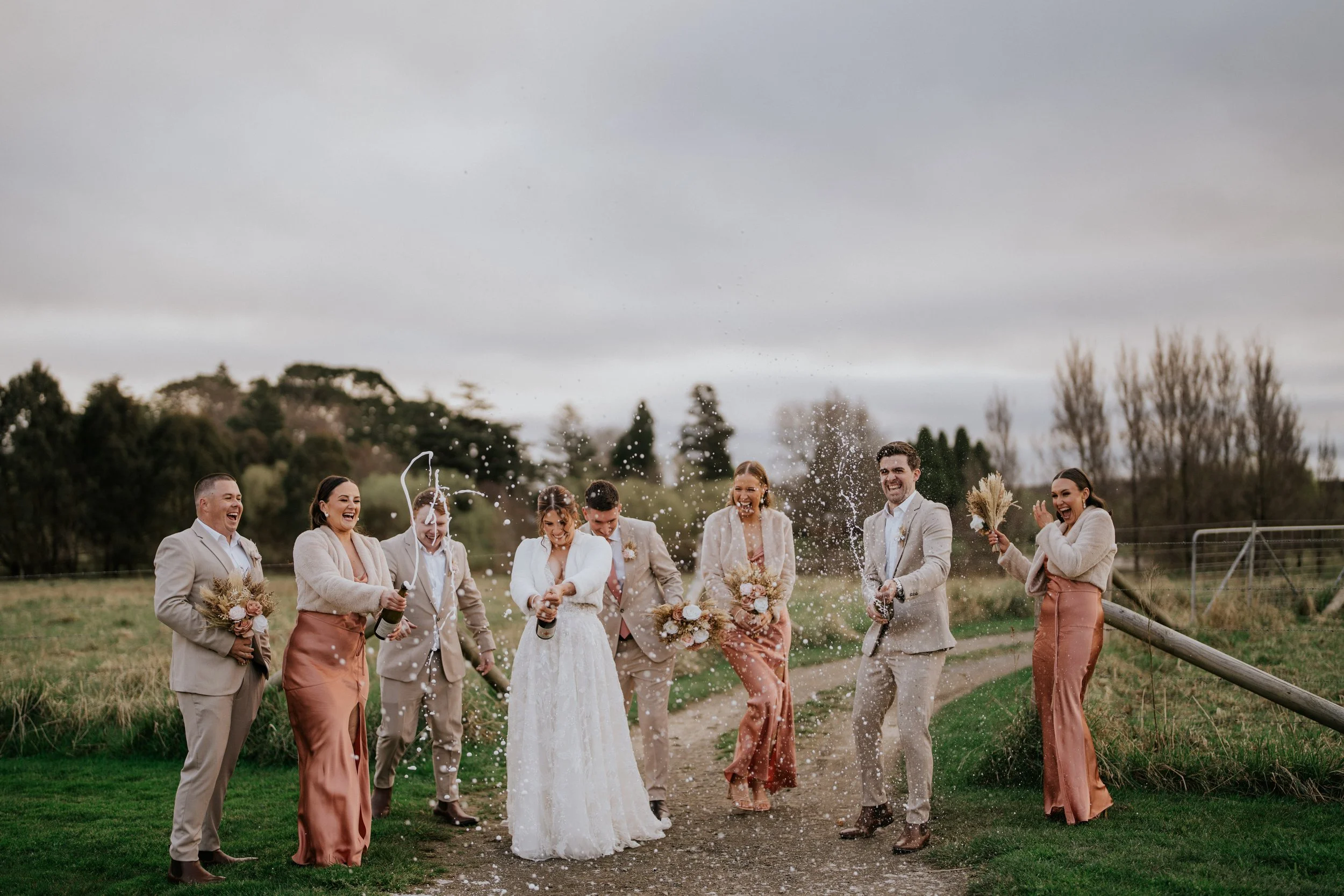 A group of wedding guests in formal attire celebrating outdoors, popping champagne and celebrating during daytime under cloudy skies.