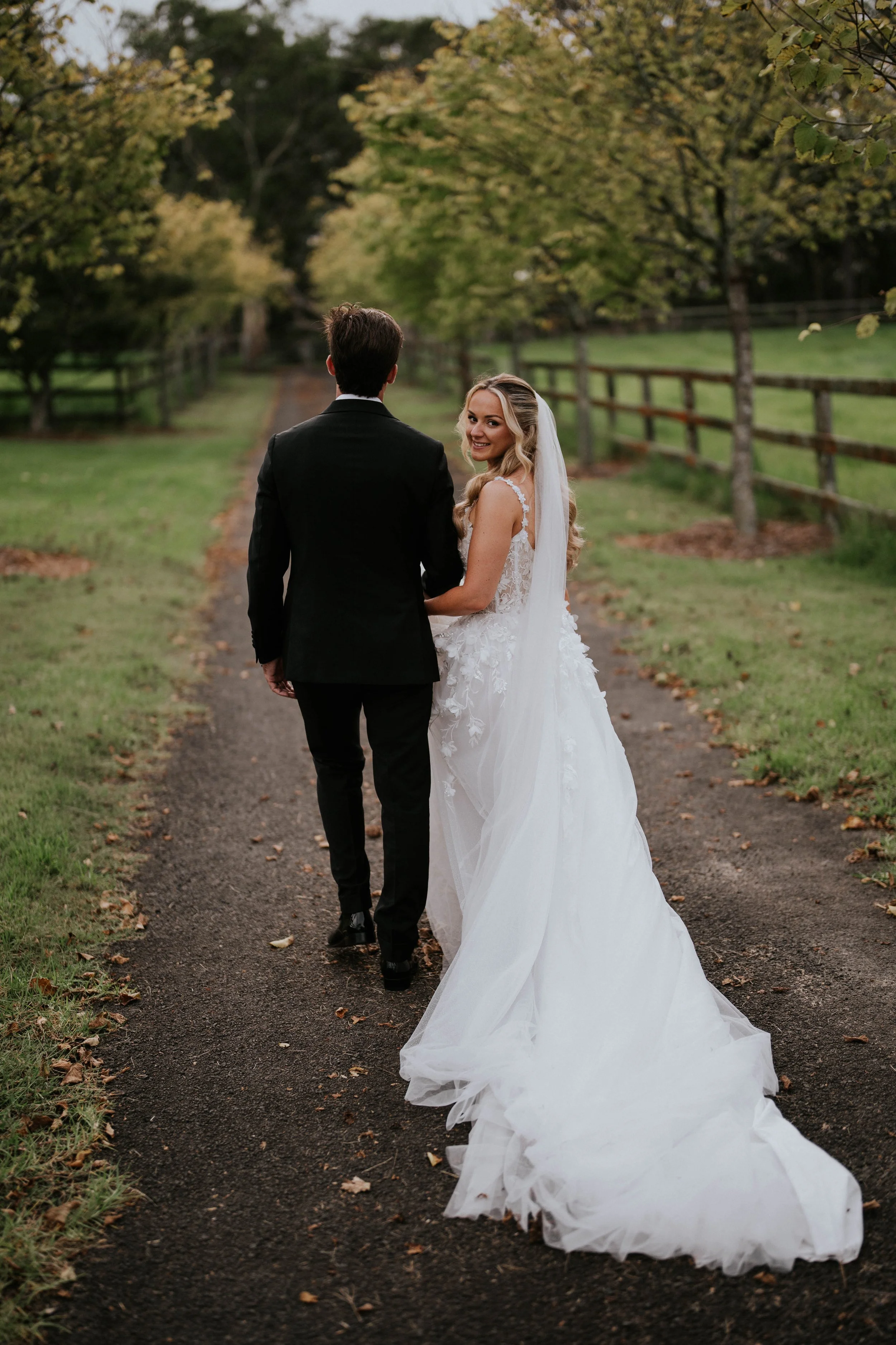 A bride and groom walking down a dirt path surrounded by trees, with the bride looking back and smiling.