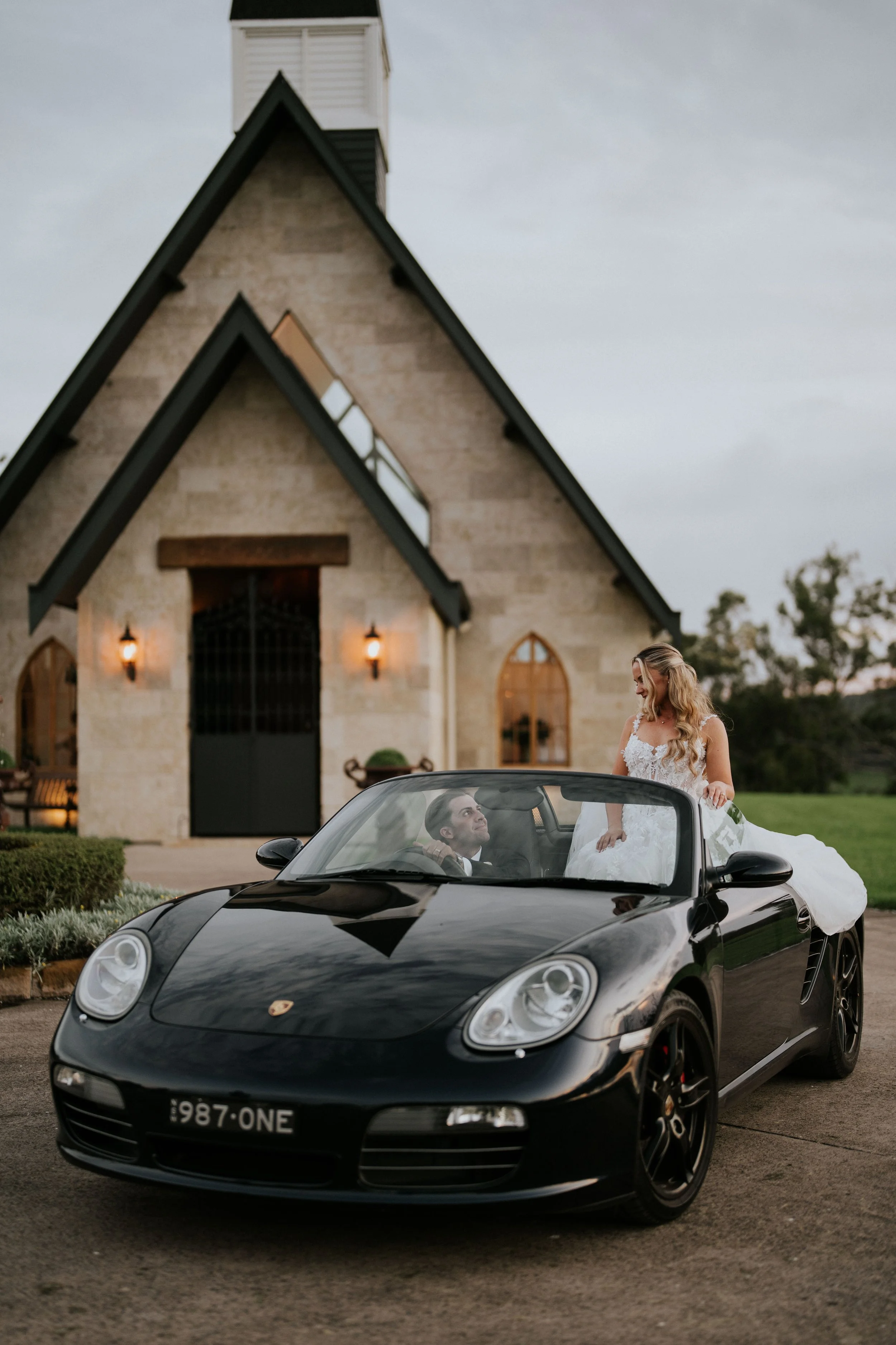 A black Porsche convertible with a bride in a white wedding dress standing outside, looking at the groom sitting in the car, in front of a church with stone walls and a peaked roof, during dusk.