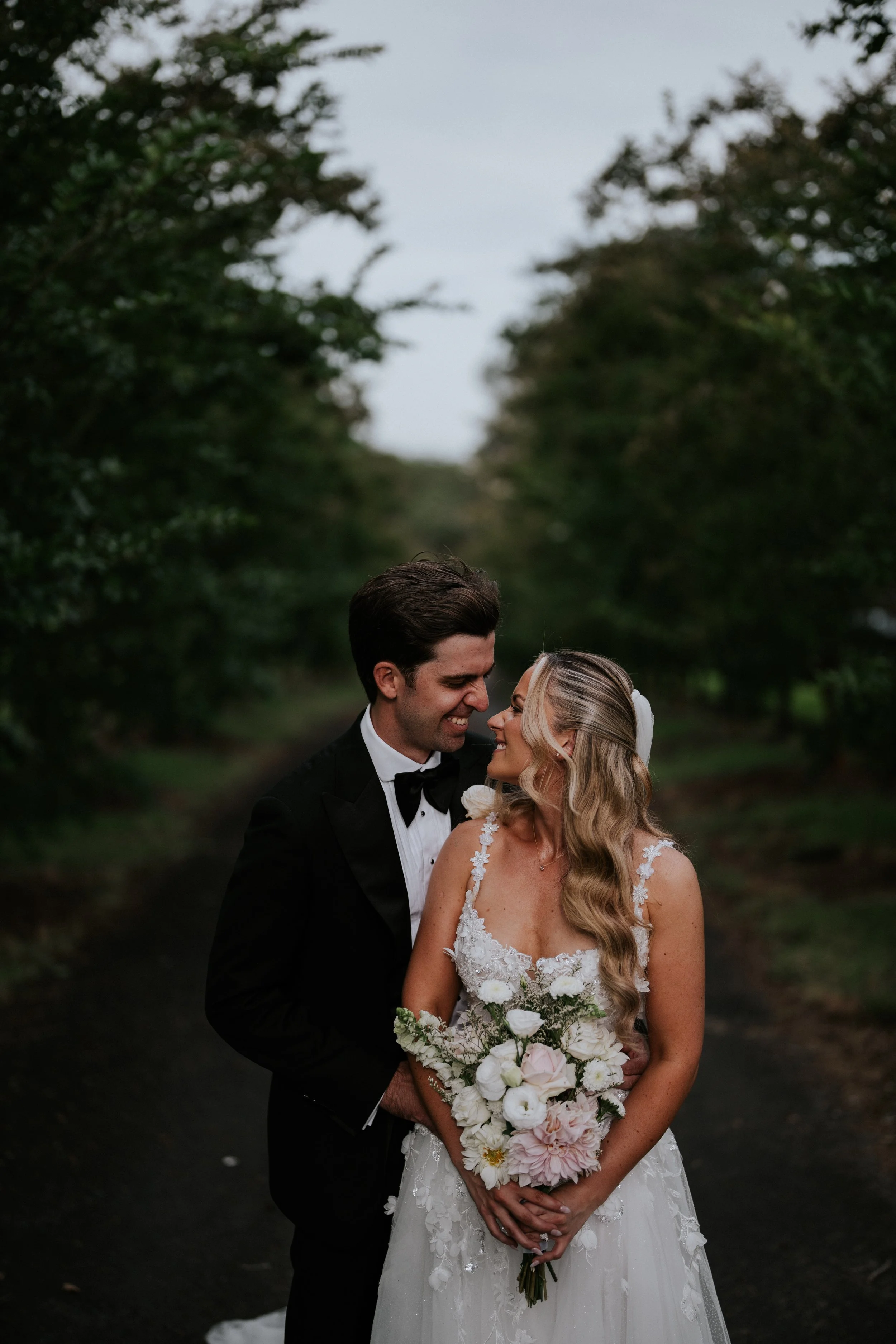 A bride and groom on their wedding day, standing close together outdoors among trees, smiling at each other. The bride holds a bouquet of white and pink flowers, and the groom is wearing a black tuxedo with a bow tie.