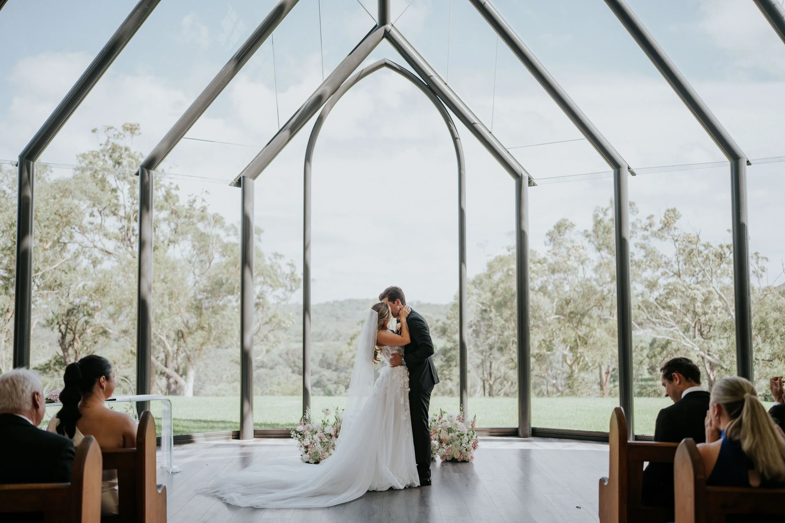 A bride and groom share a kiss at their wedding ceremony inside a glass chapel with an outdoor landscape in the background.