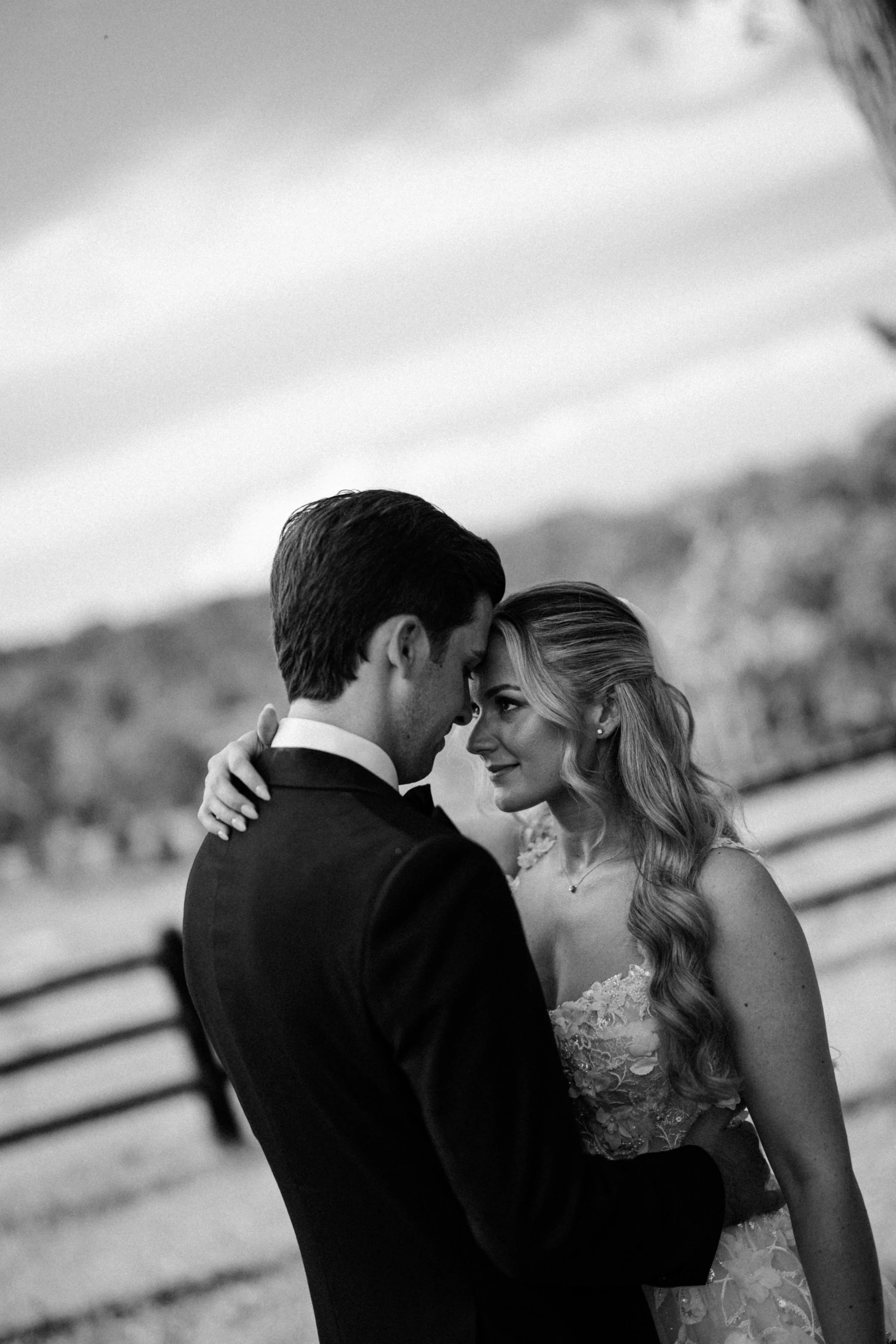 A black and white photo of a couple in wedding attire, embracing and looking into each other's eyes outdoors, with a blurred landscape in the background.
