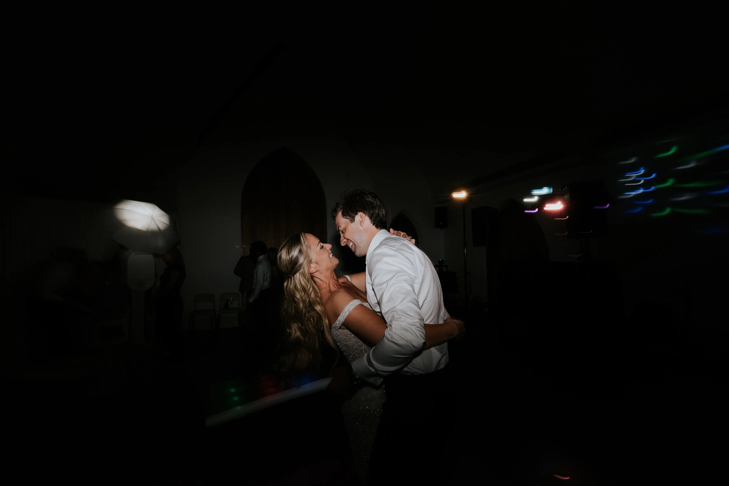 A couple dancing closely at a celebration or wedding reception, smiling and looking into each other's eyes in a dimly lit room with colorful lights in the background.