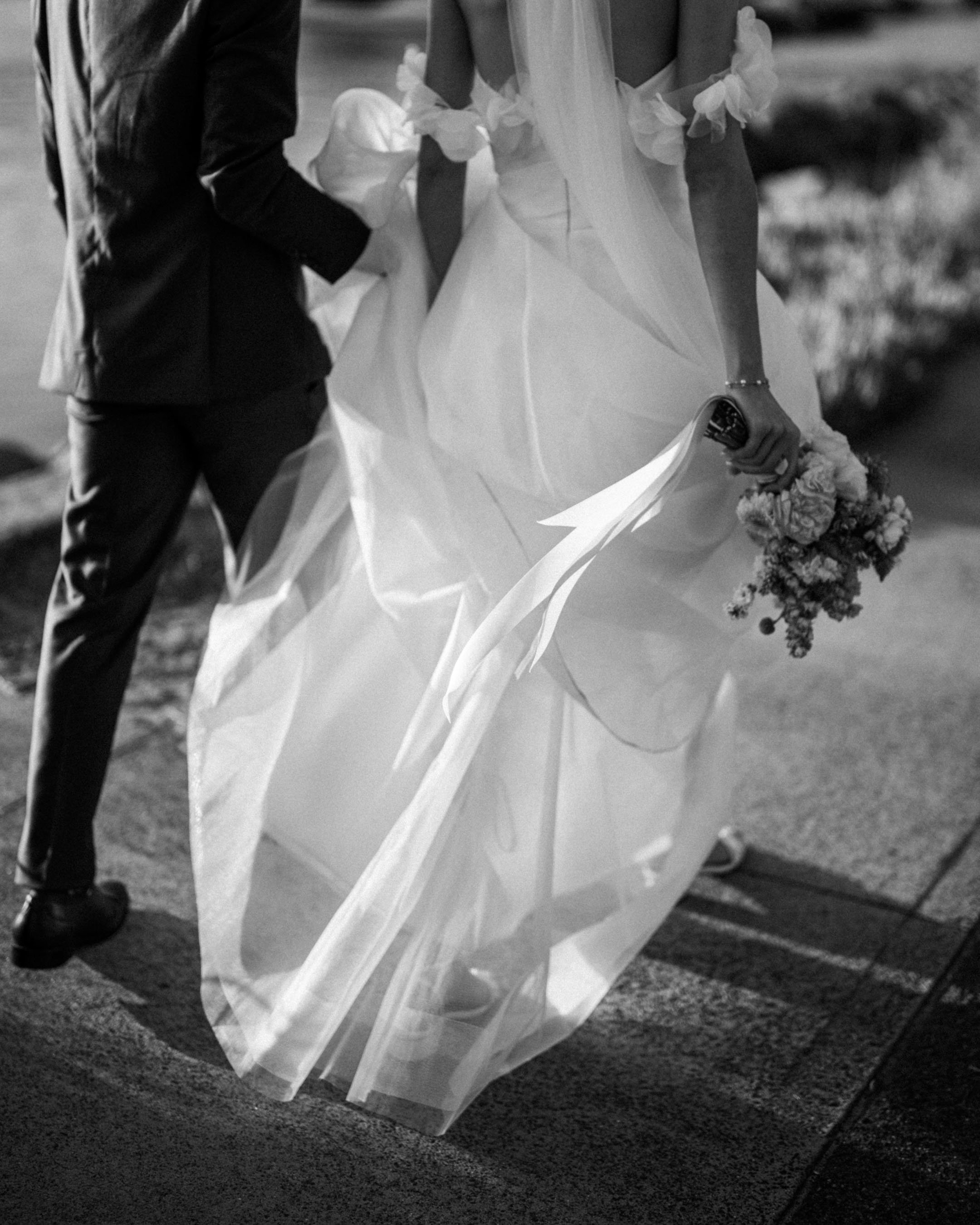 Black and white photo of a bride and groom walking outdoors; the bride is holding a bouquet of flowers, and the groom is dressed in a suit, holding her hand.