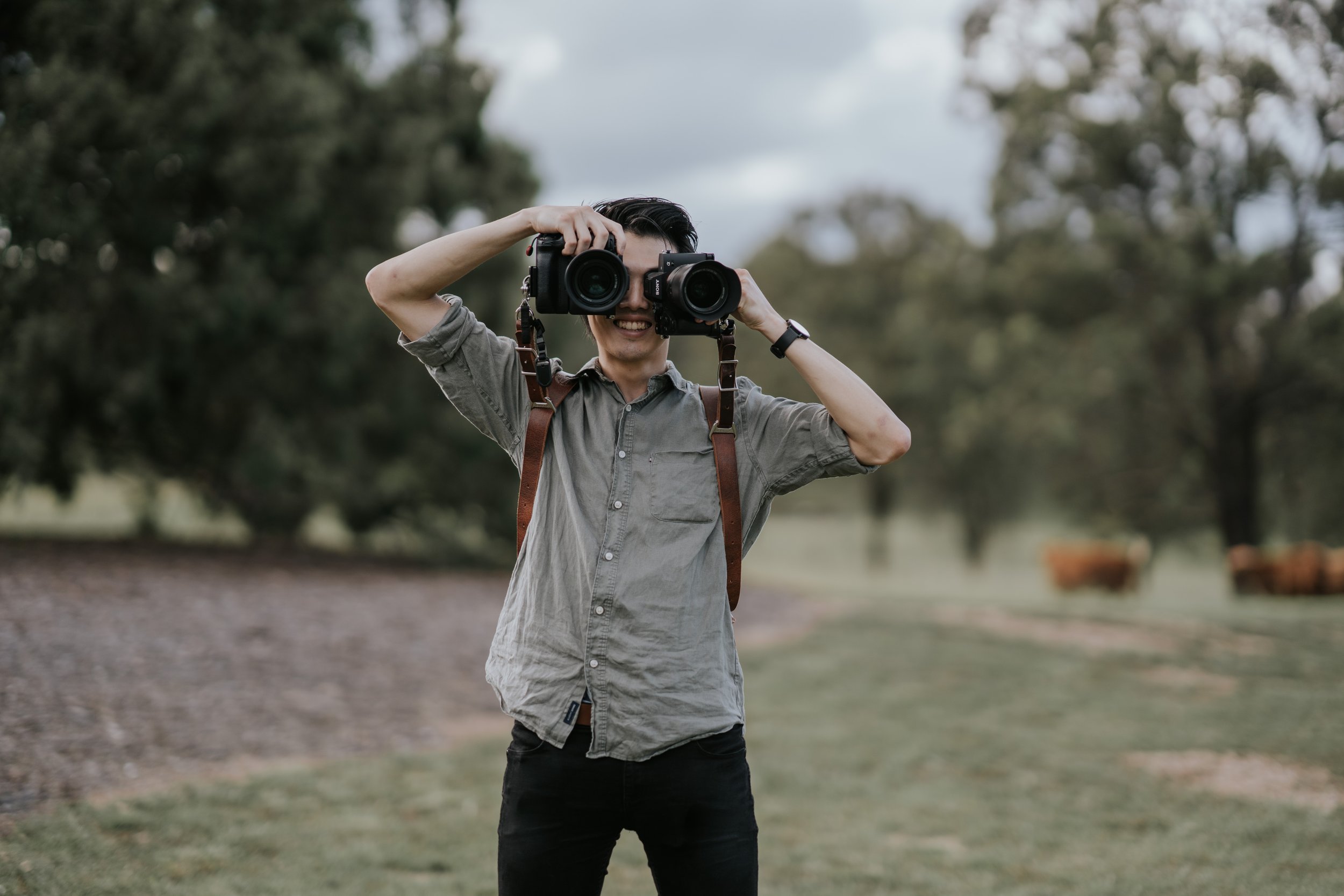 Person smiling and holding two cameras up to their face outdoors in a park with trees in the background.