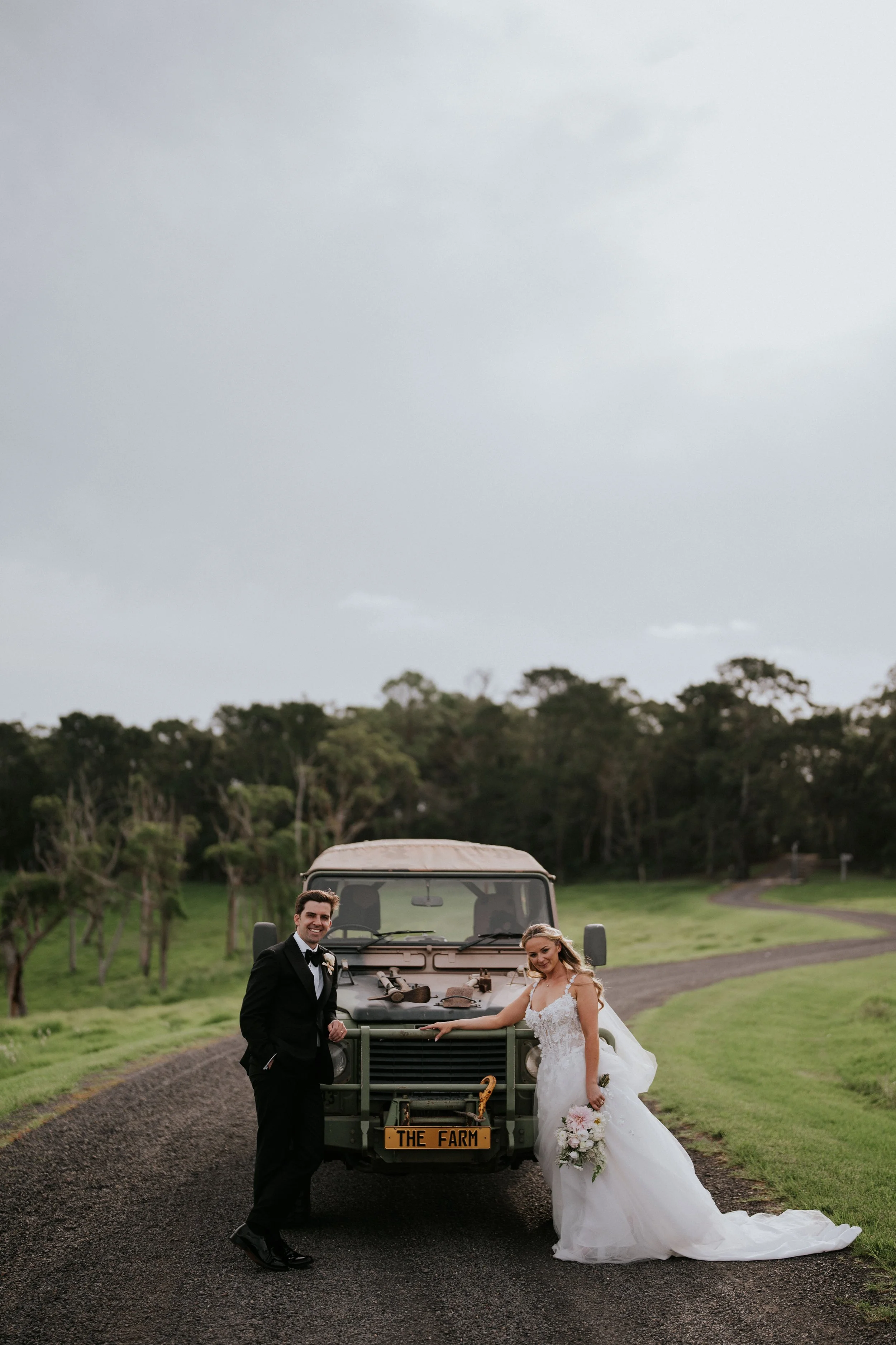A bride and groom in wedding attire standing in front of a vintage military vehicle on a farm, holding hands and smiling, with trees and a winding road in the background.