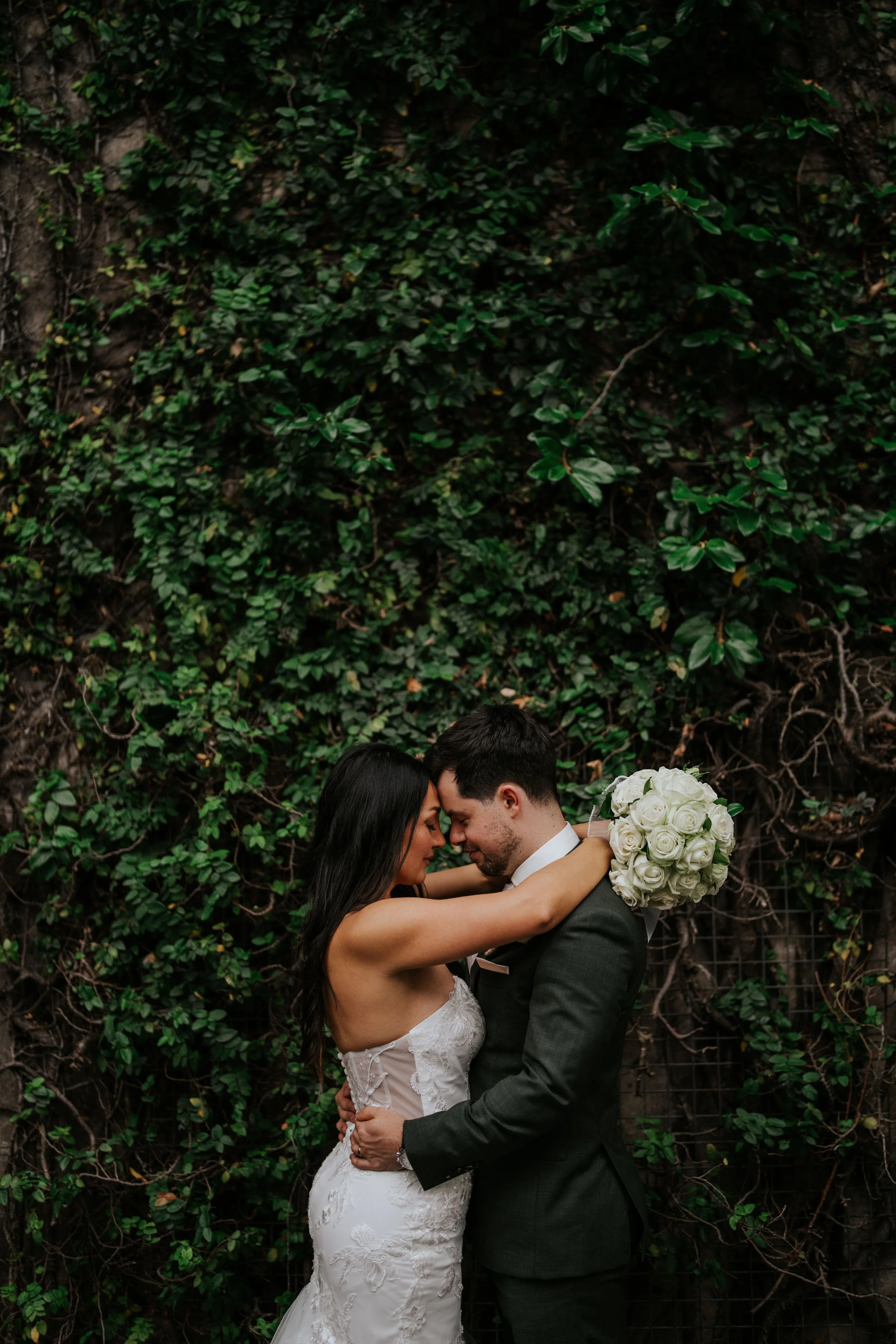 A bride and groom are embracing and touching foreheads with eyes closed, standing in front of a green ivy-covered wall. The bride is wearing a white lace wedding dress and the groom is in a dark suit. The bride is holding a bouquet of white roses.