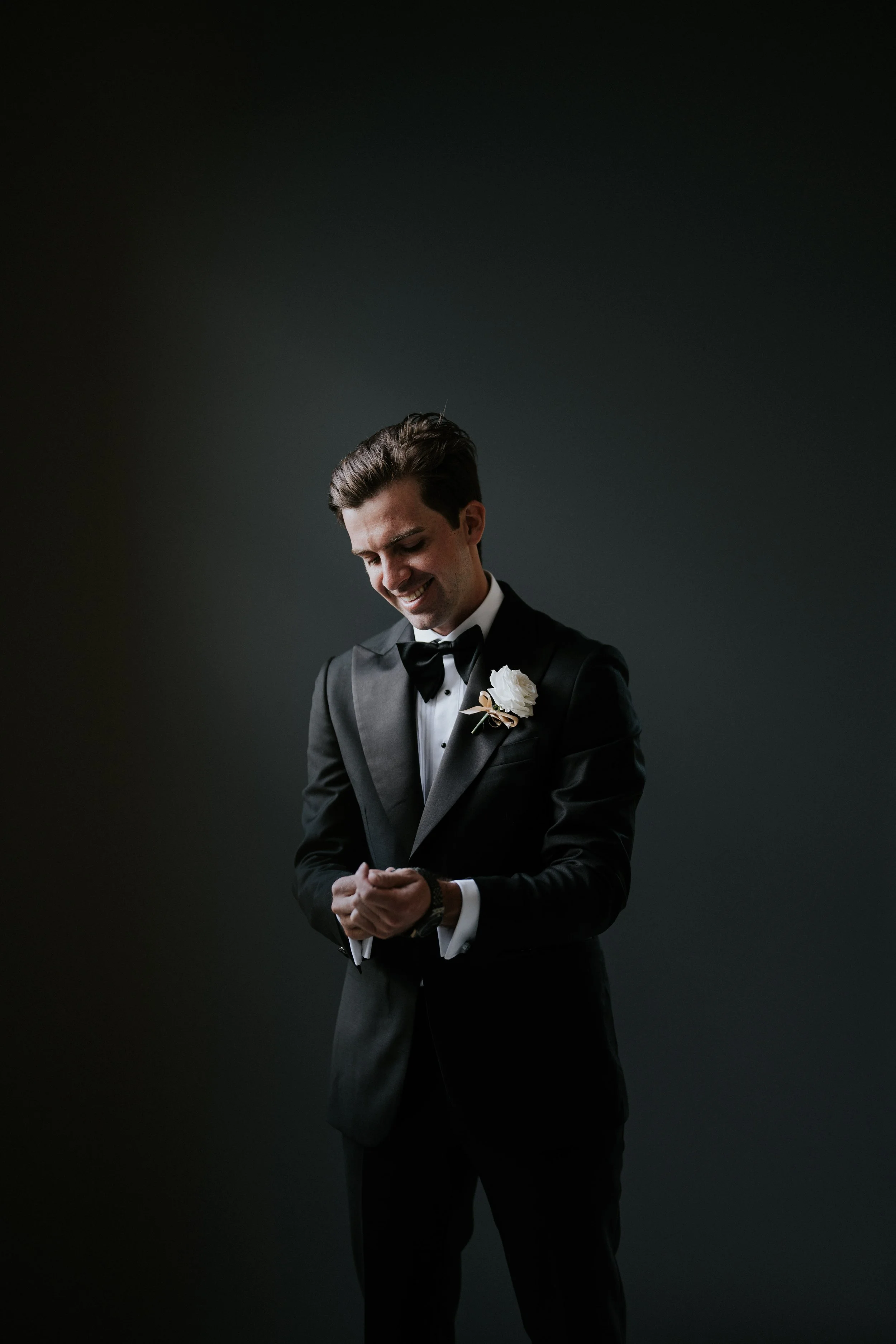 A smiling man in a black tuxedo with a white boutonniere is standing against a dark background, looking down with his hands clasped.