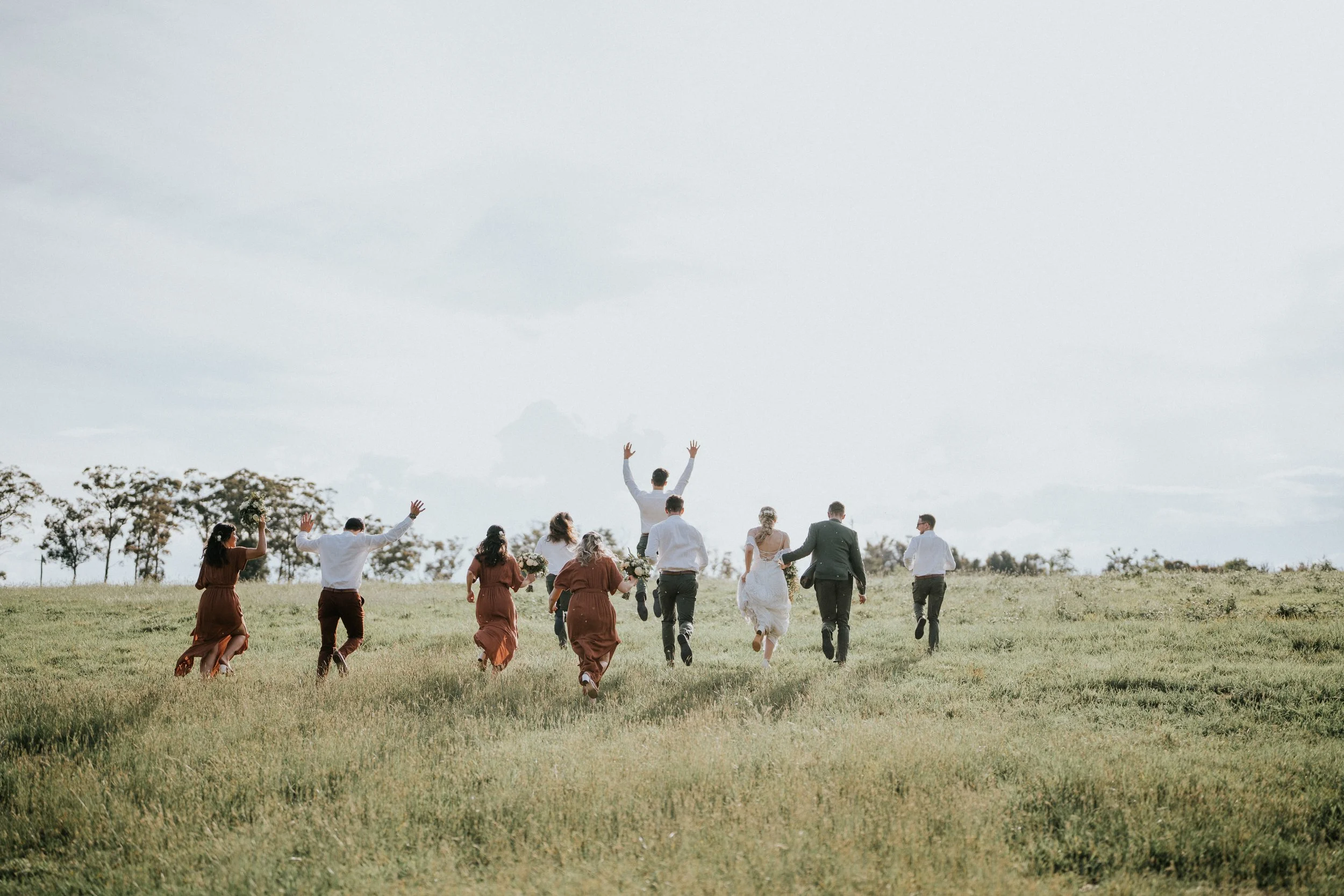 A group of people, including a bride in a white dress, walking away from the camera across a grassy field in an outdoor wedding ceremony, with a group of bridesmaids in rust-colored dresses and groomsmen in suits, and a person raising their arms in the air.