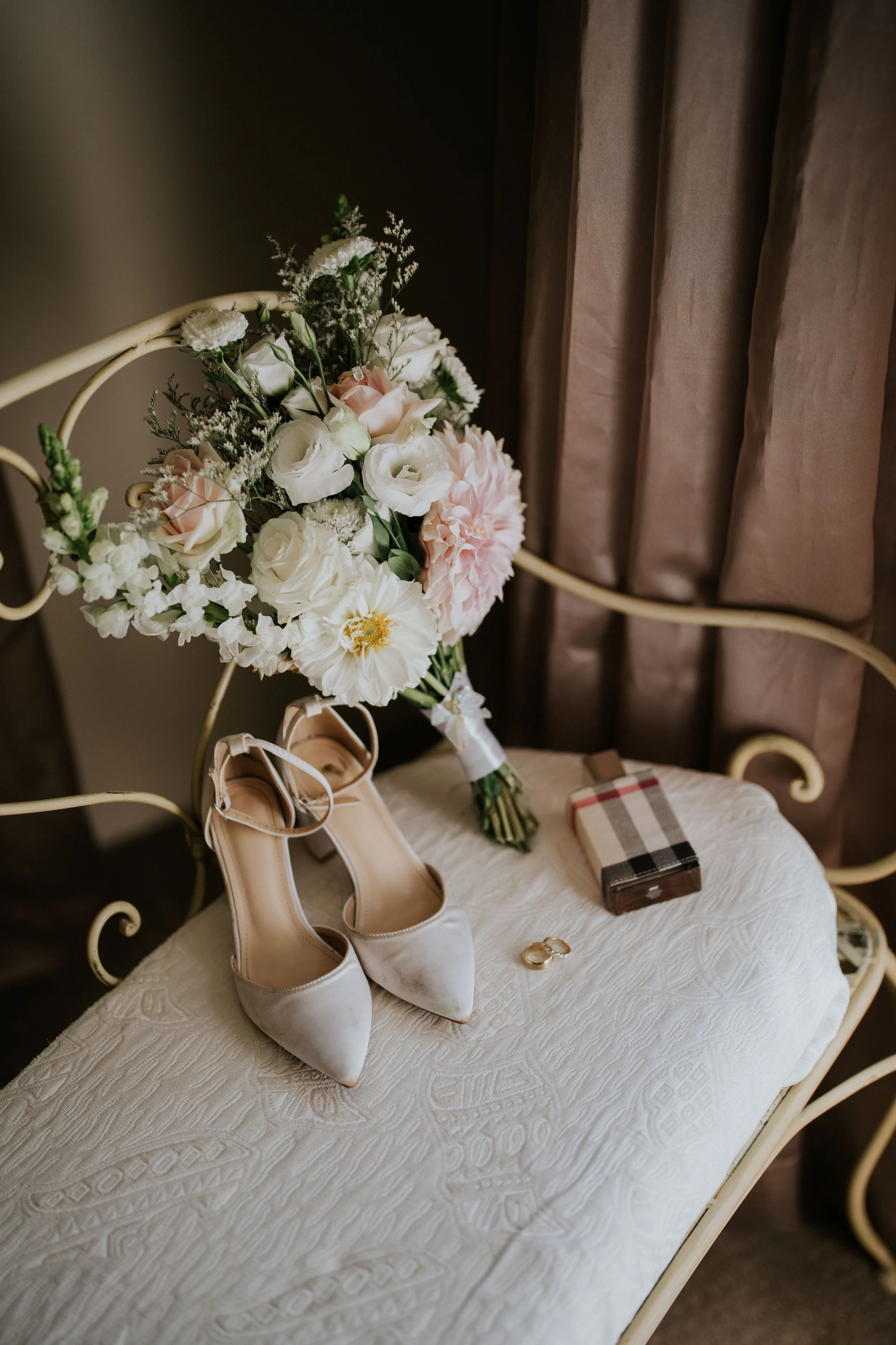 A bench with a bouquet of white and pink flowers, a pair of white bridal shoes, jewelry, a wrapped gift, and a patterned box on it, against a background of beige curtains.