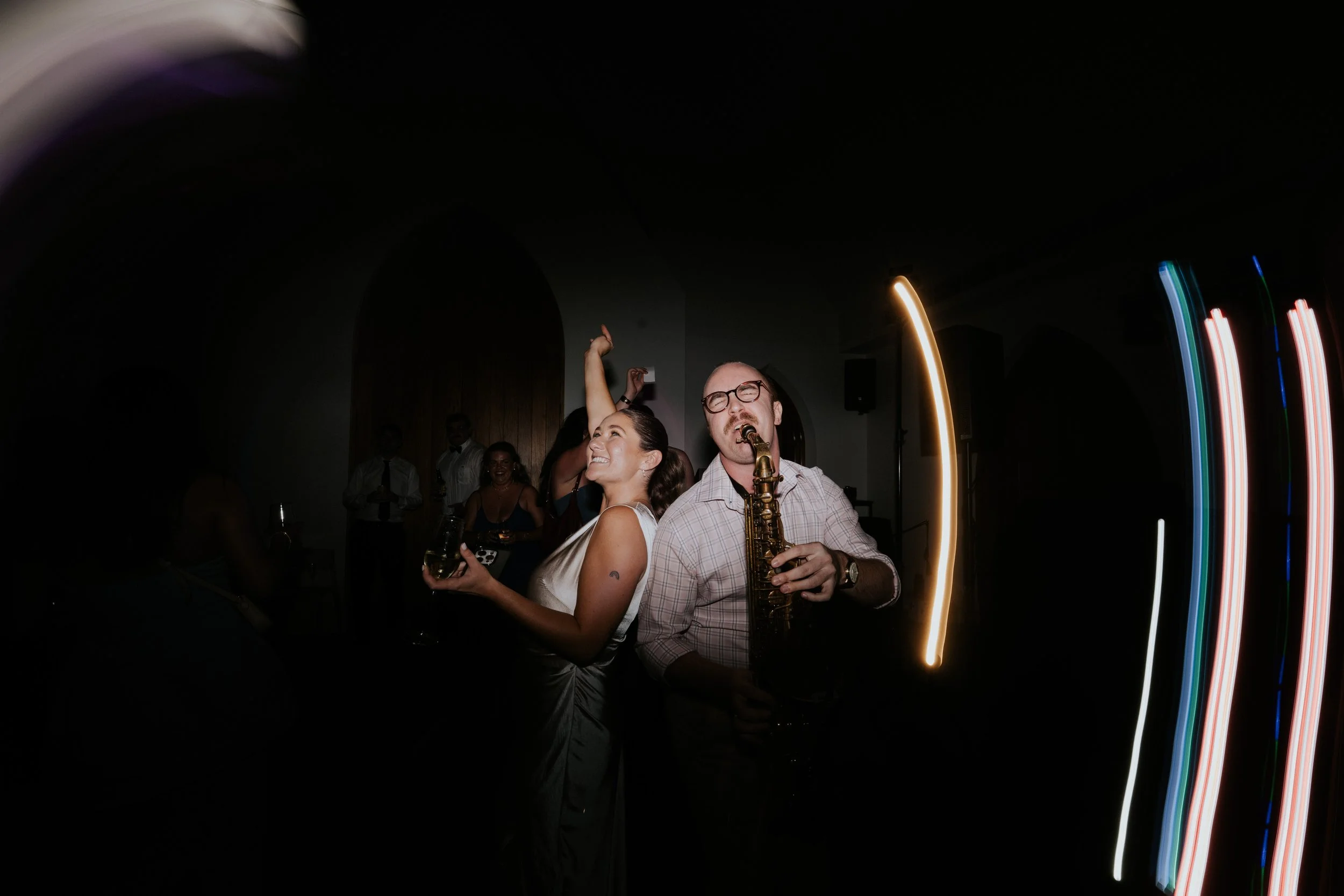 A lively indoor party scene with a man playing a saxophone and a smiling woman holding a glass of wine, surrounded by other guests, with colorful light streaks in the background.