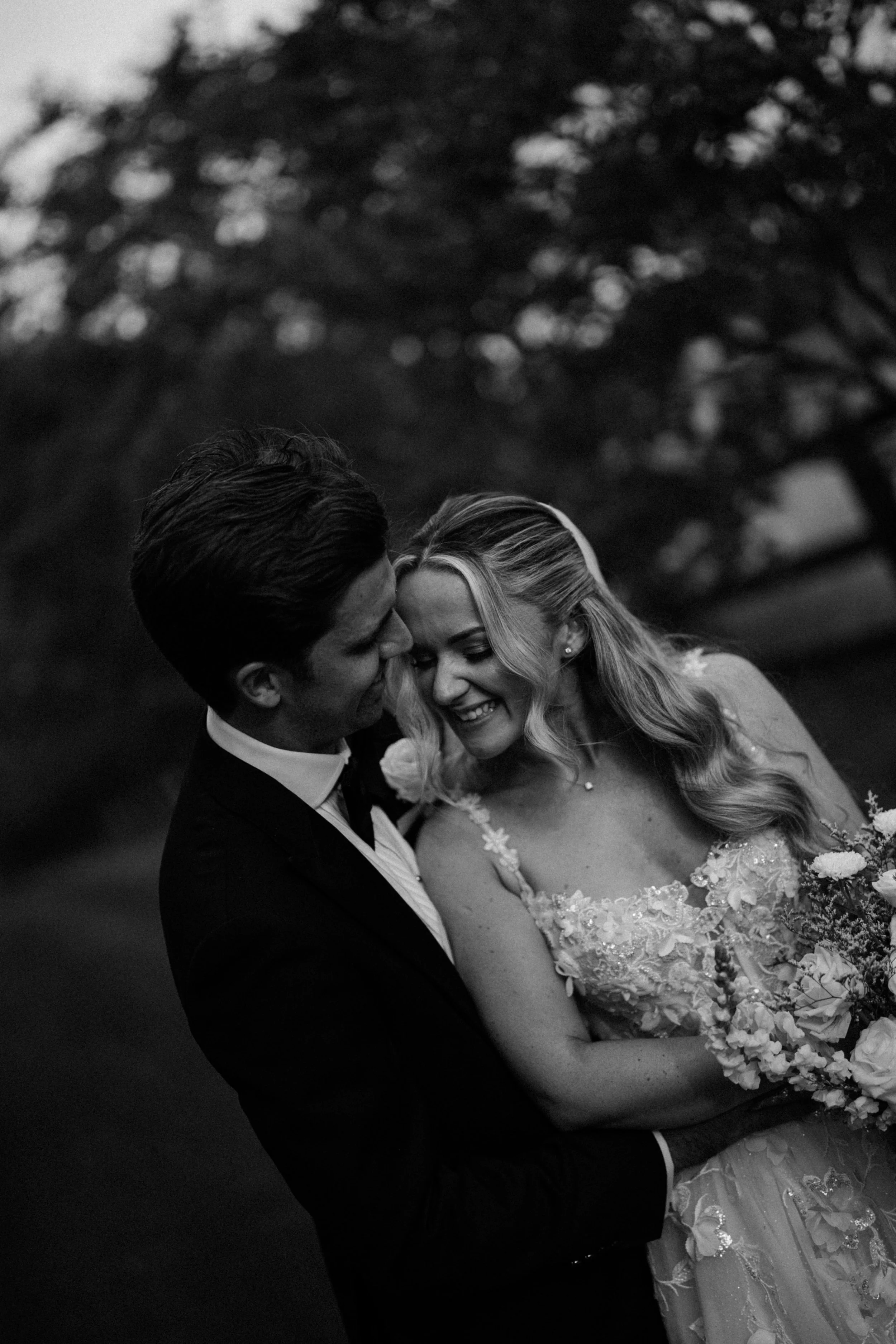 Black and white photo of a bride and groom leaning close together, smiling and touching foreheads, outdoors with a blurred background of trees.