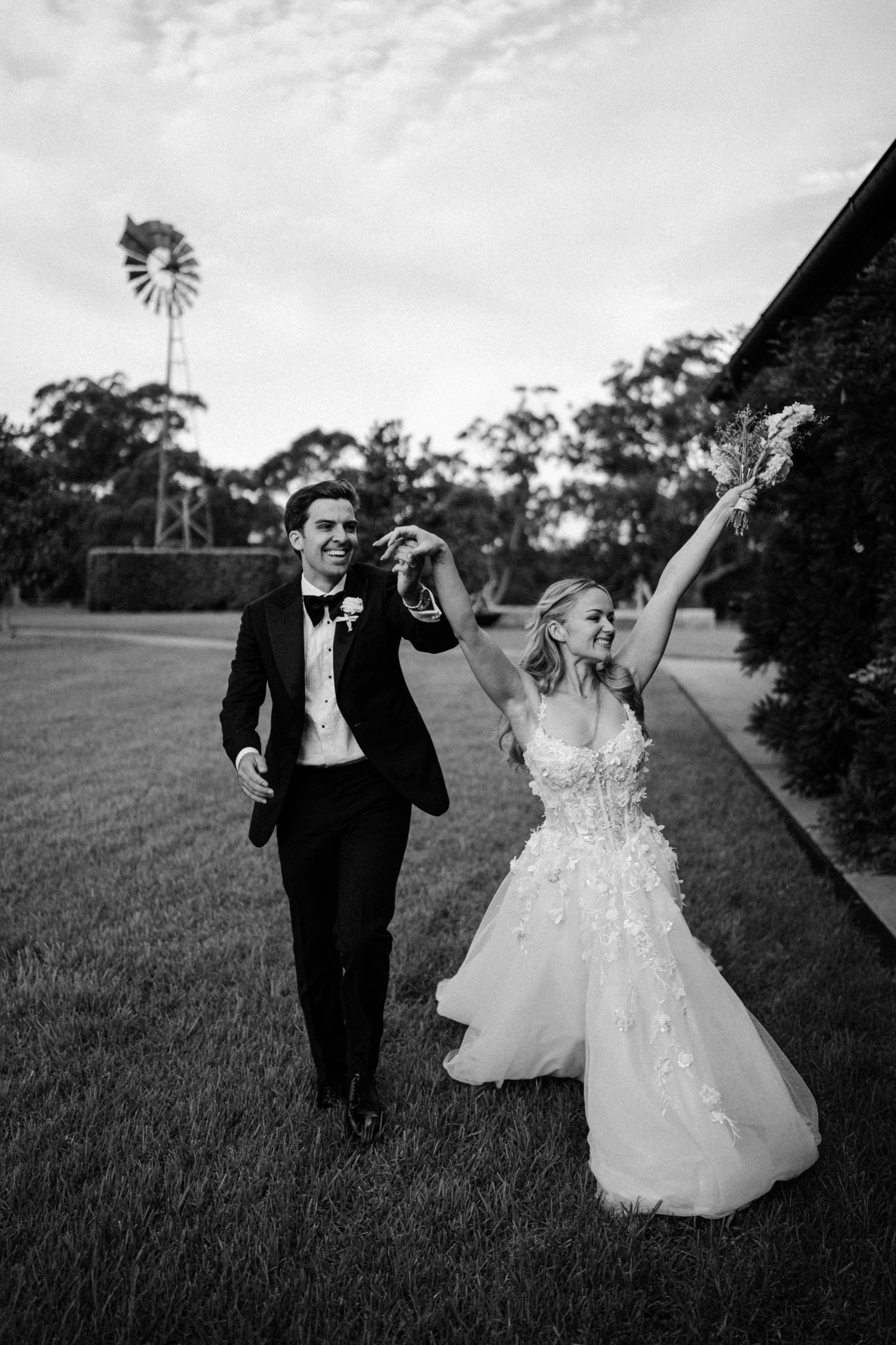 A black and white photo of a bride and groom dancing outdoors on a grassy area. The bride is holding a bouquet with one arm raised, and the groom is smiling, holding her hand gently. There is a windmill and trees in the background.