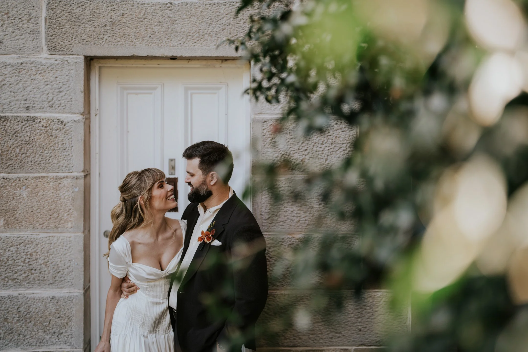 A couple dressed in wedding attire standing close to each other, smiling, outside near a textured beige stone building with a white door, partially obscured by greenery.