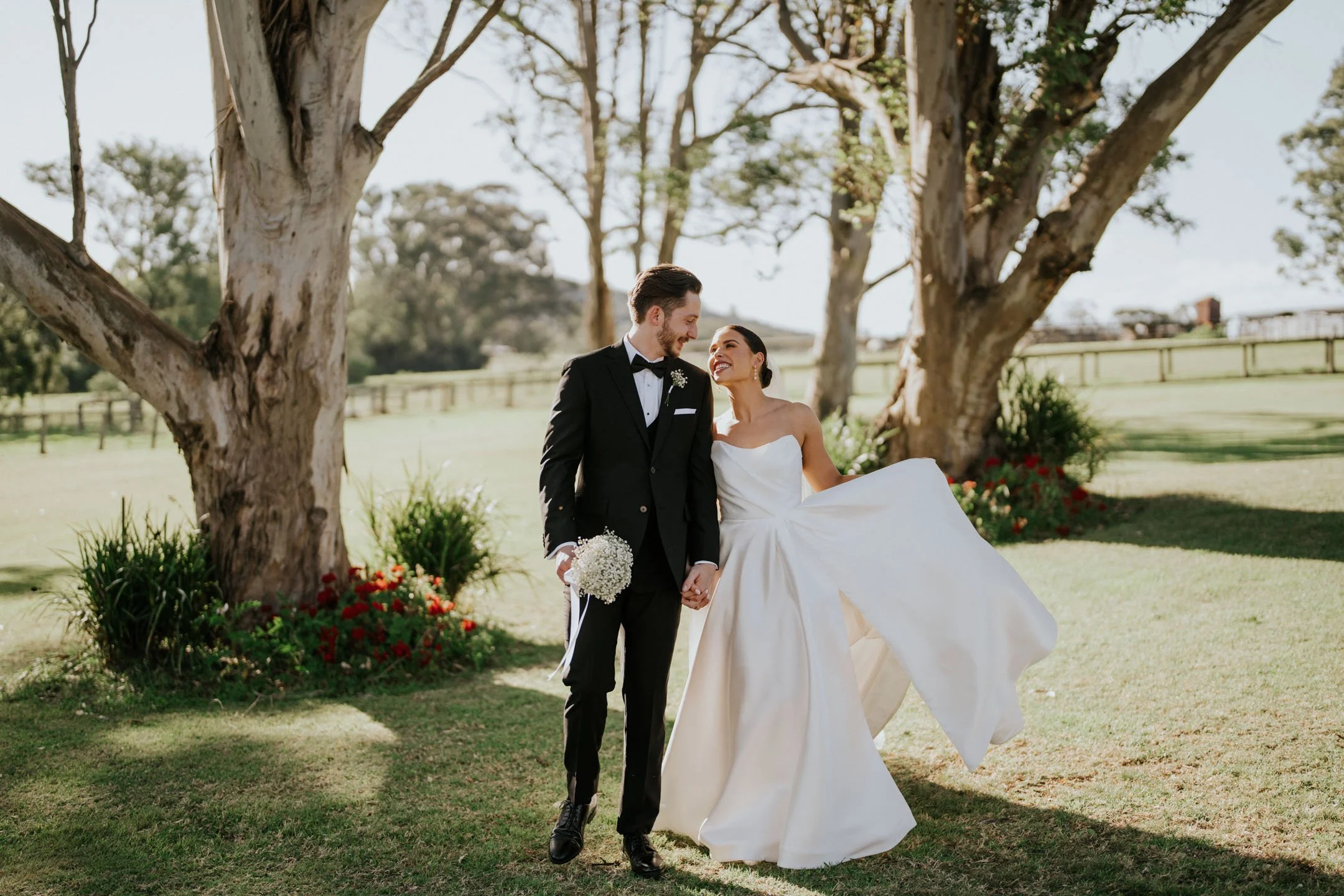 A bride and groom dressed in wedding attire walking hand in hand outdoors near trees, smiling at each other during their wedding celebration.