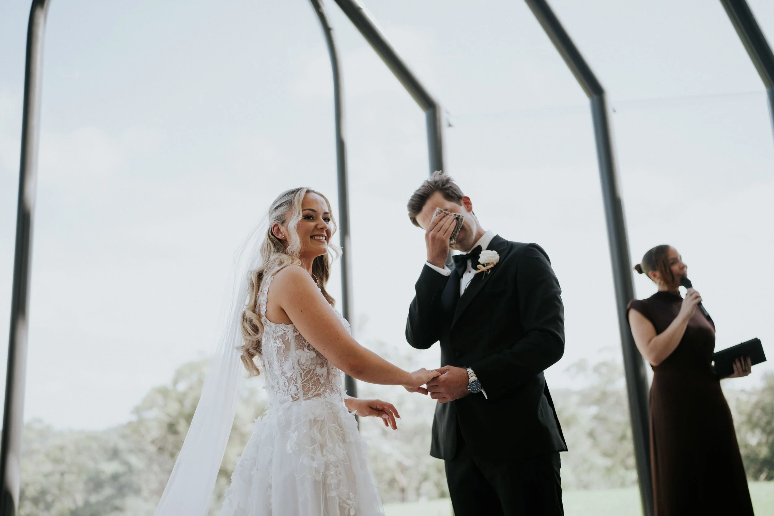 A bride and groom standing outdoors holding hands during their wedding ceremony, with the groom wiping his eyes with a tissue, and a woman speaking into a microphone in the background.