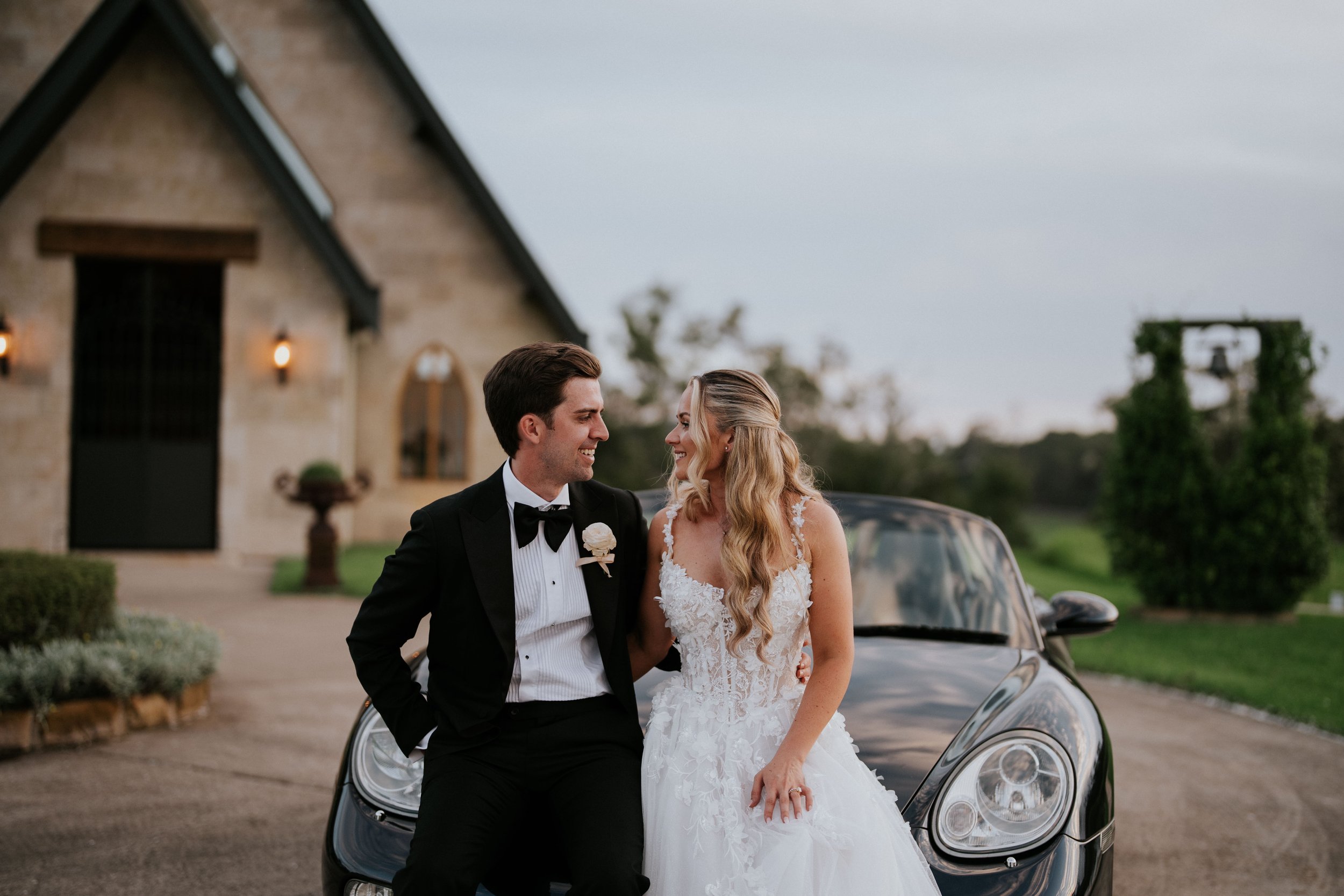 A bride and groom in wedding attire standing in front of a black car, smiling at each other near a rustic building with lights and greenery in the background.
