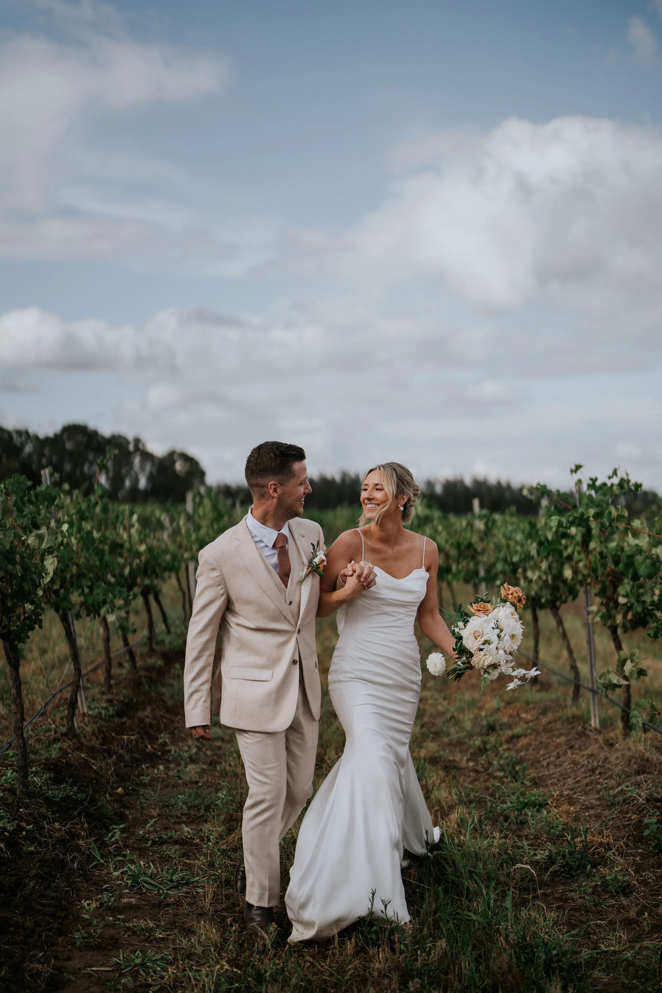 A bride and groom walking together through a vineyard on a cloudy day, holding hands and smiling.