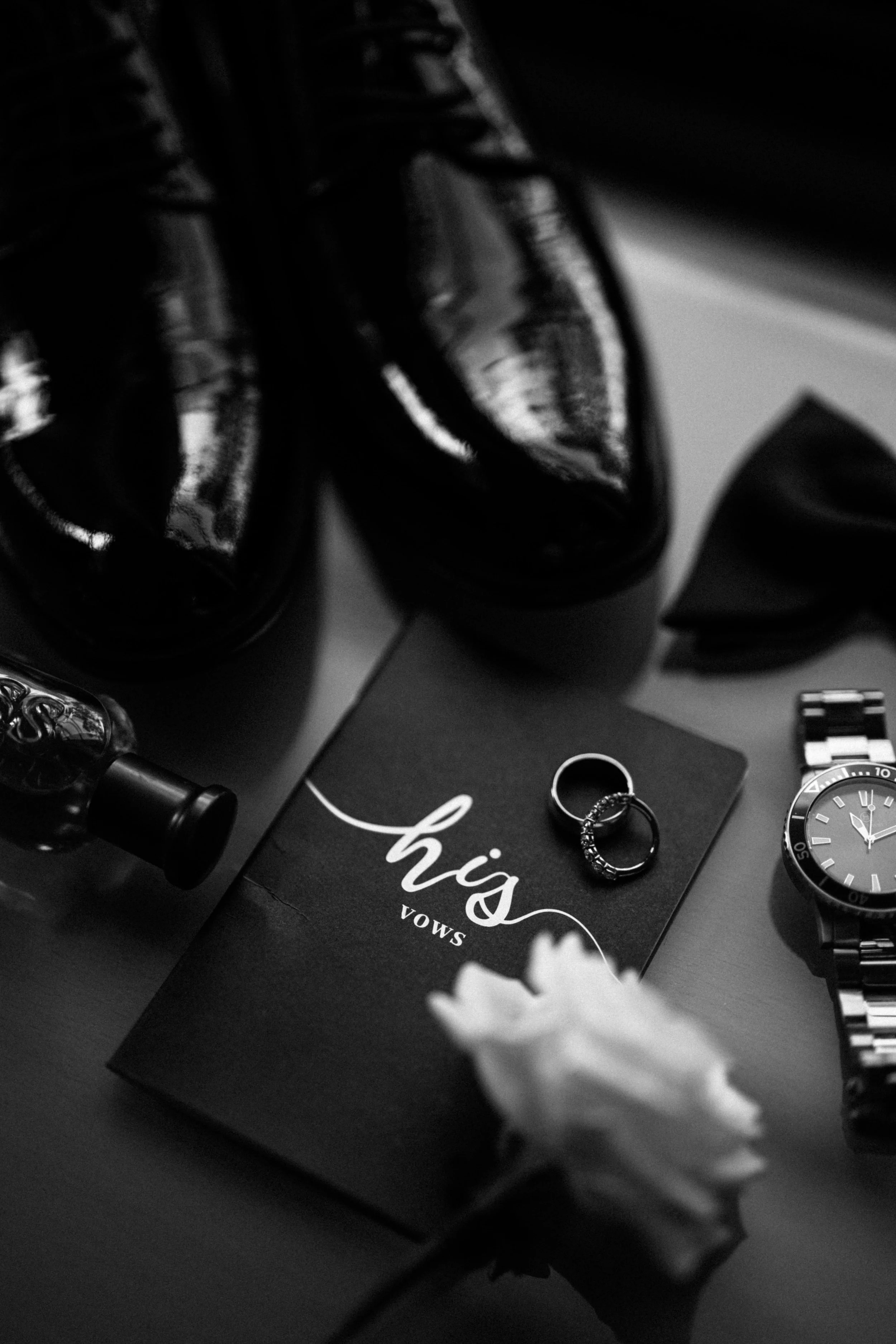 Black leather shoes, a perfume bottle, jewelry including rings, a watch, and a gift box with a white flower on top.