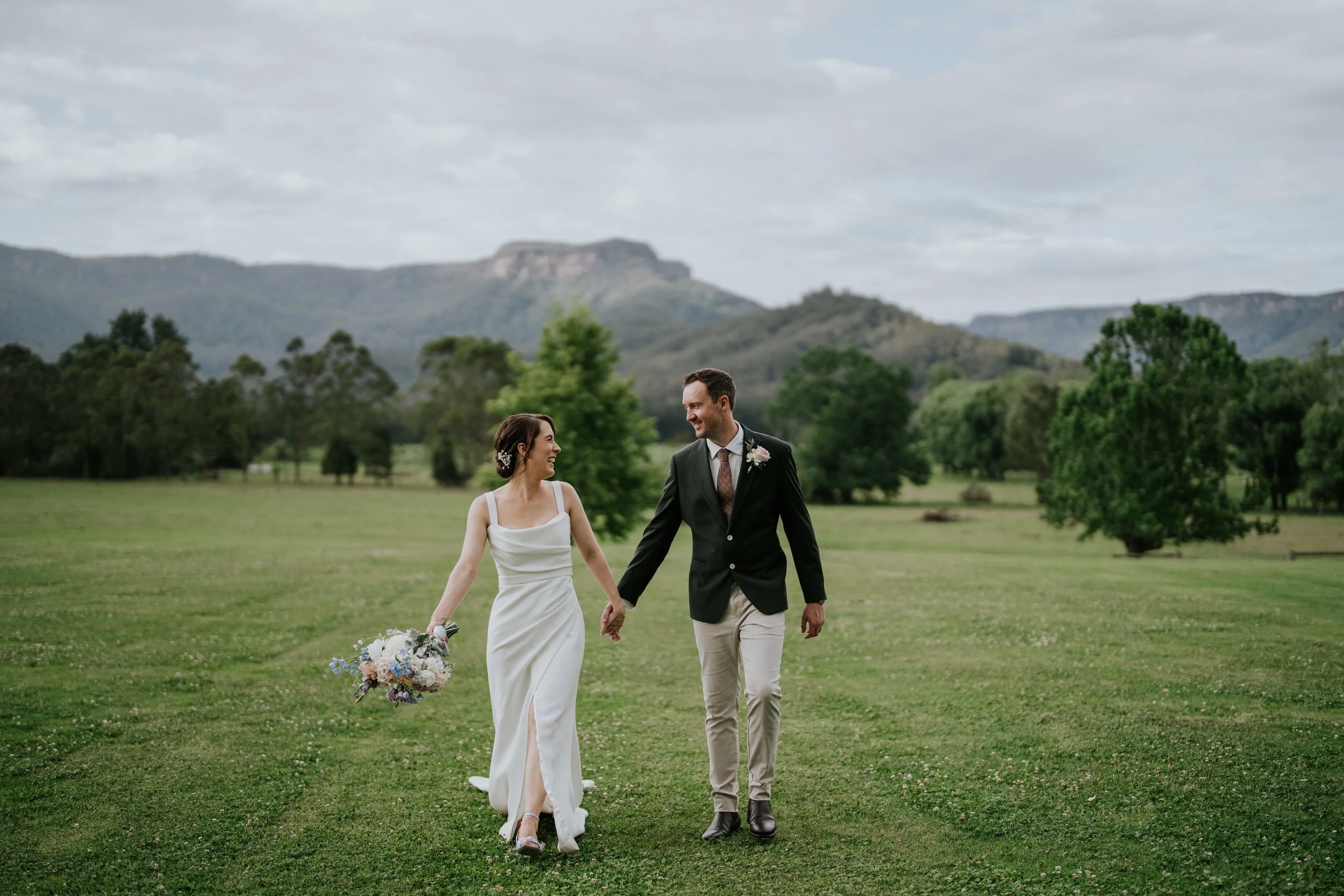 A bride and groom walking hand in hand on a grassy field, with the bride holding a bouquet of flowers, and mountains and trees in the background on a cloudy day.