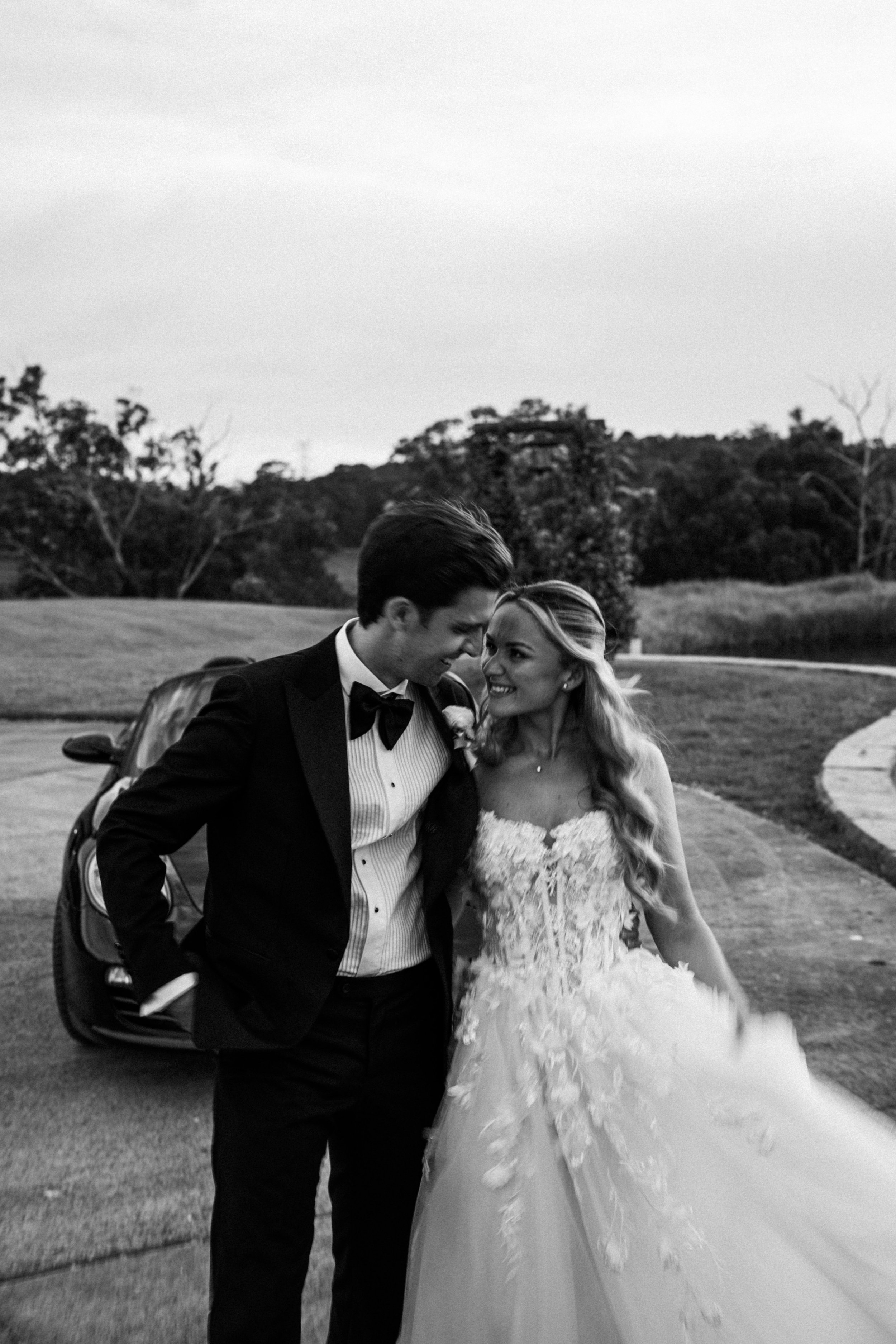 A newlywed couple smiling and gazing at each other outdoors in a park, with a car in the background and trees around them, black and white photo.