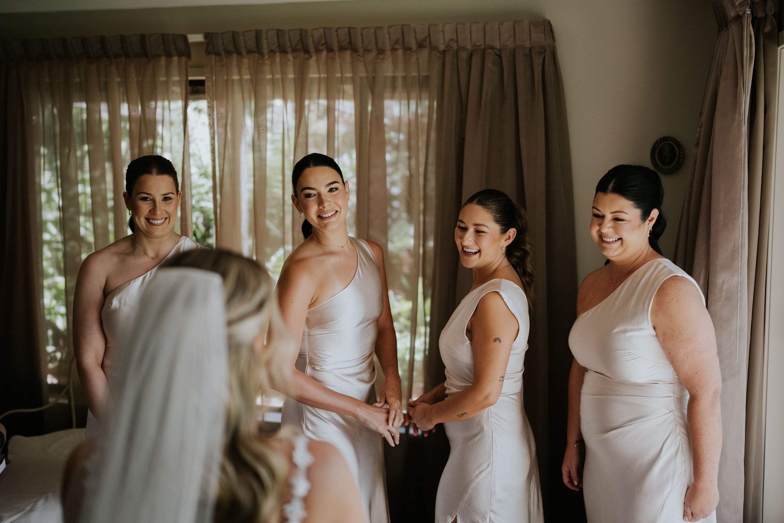 Four women in white dresses holding hands and smiling in front of a mirror, surrounded by curtains in a bright room.